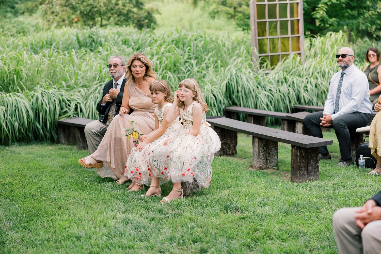 Wedding guests relaxing on wooden benches in a lush garden setting during an outdoor reception.