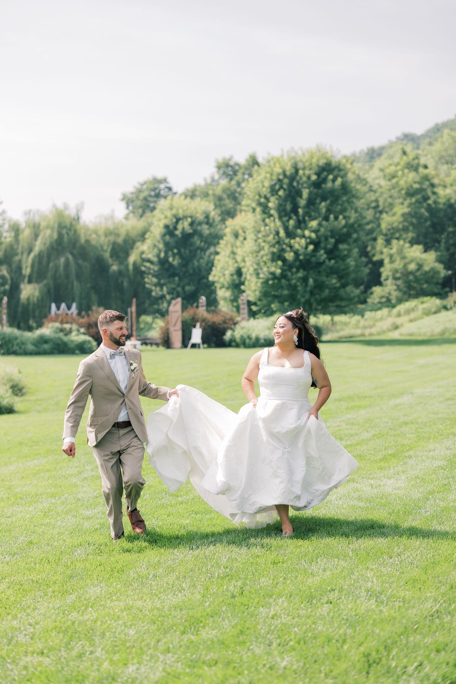 A couple runs playfully across a sunny green field with flowing white fabric billowing in the wind.