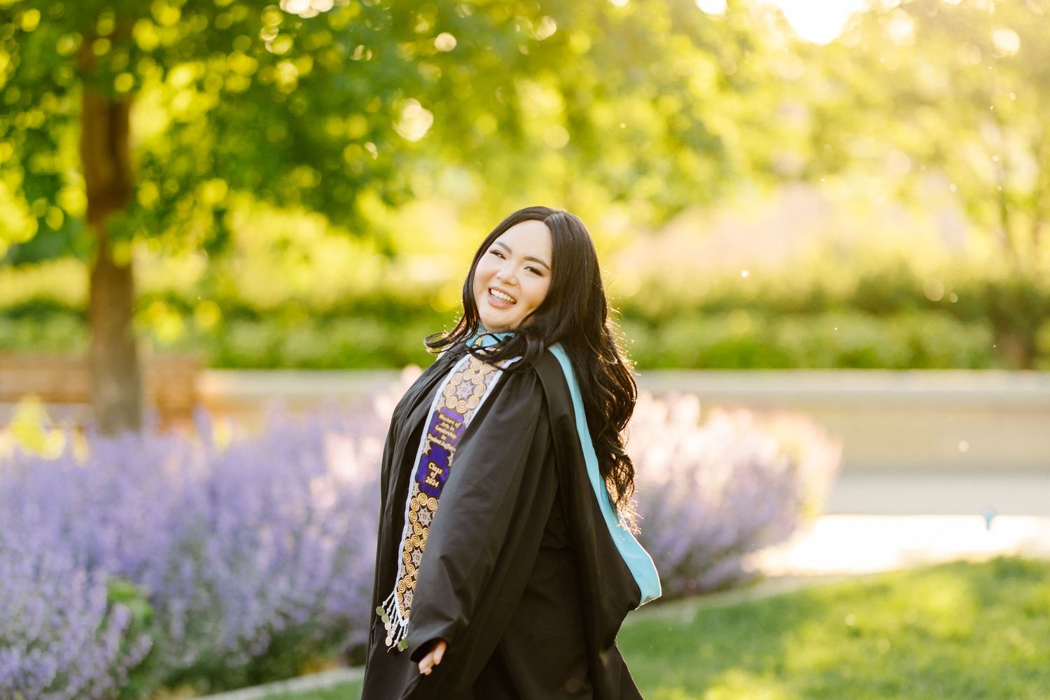 Graduate in academic dress twirls joyfully in front of blooming lavender garden during sunset.