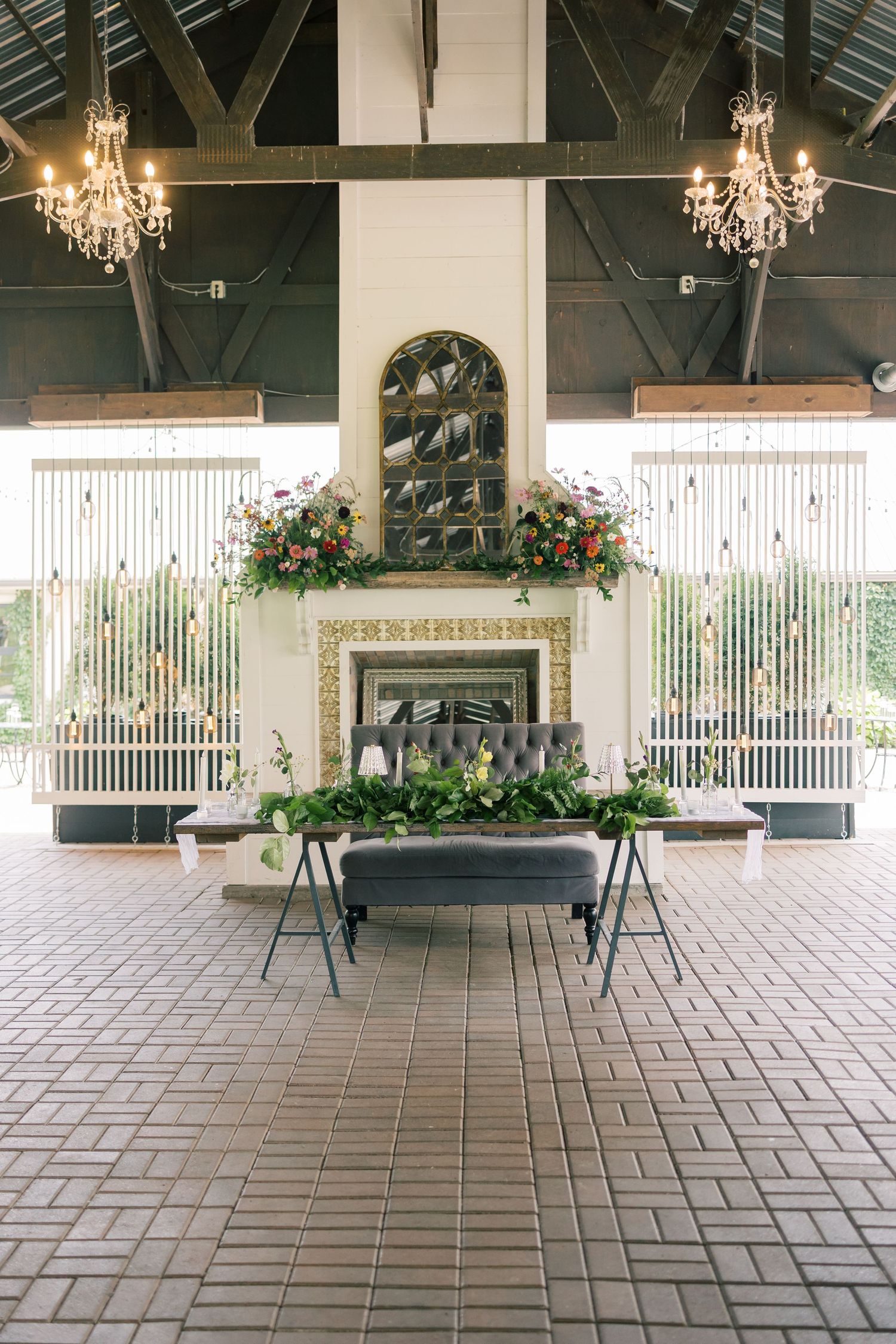 Elegant covered outdoor venue with brick flooring, crystal chandeliers, and white columns creating an upscale event space.
