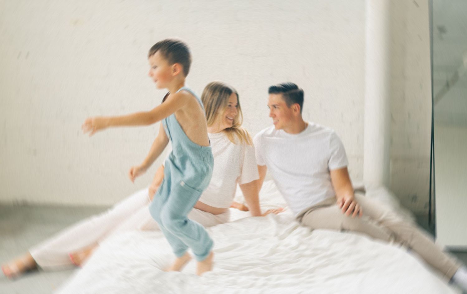 A happy family with a young boy jumping on a white bed while parents smile in a bright, airy room.