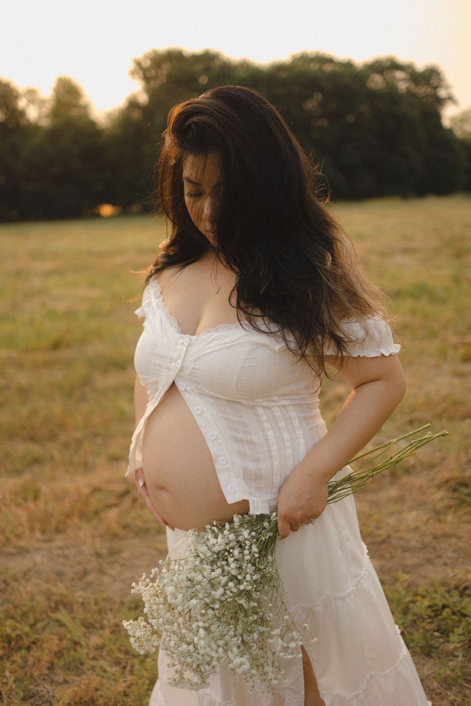 Pregnant woman in white dress holding wildflowers in a golden sunset field, cradling her bare baby bump.