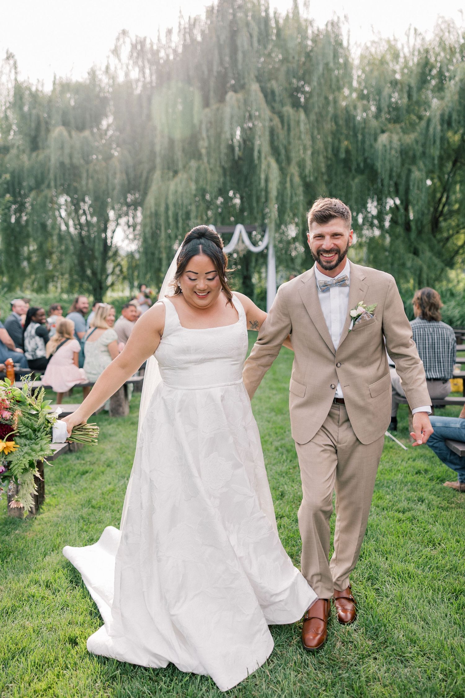 A wedding couple shares playful moments outdoors, walking and dancing together in beige and white attire on a grassy lawn.