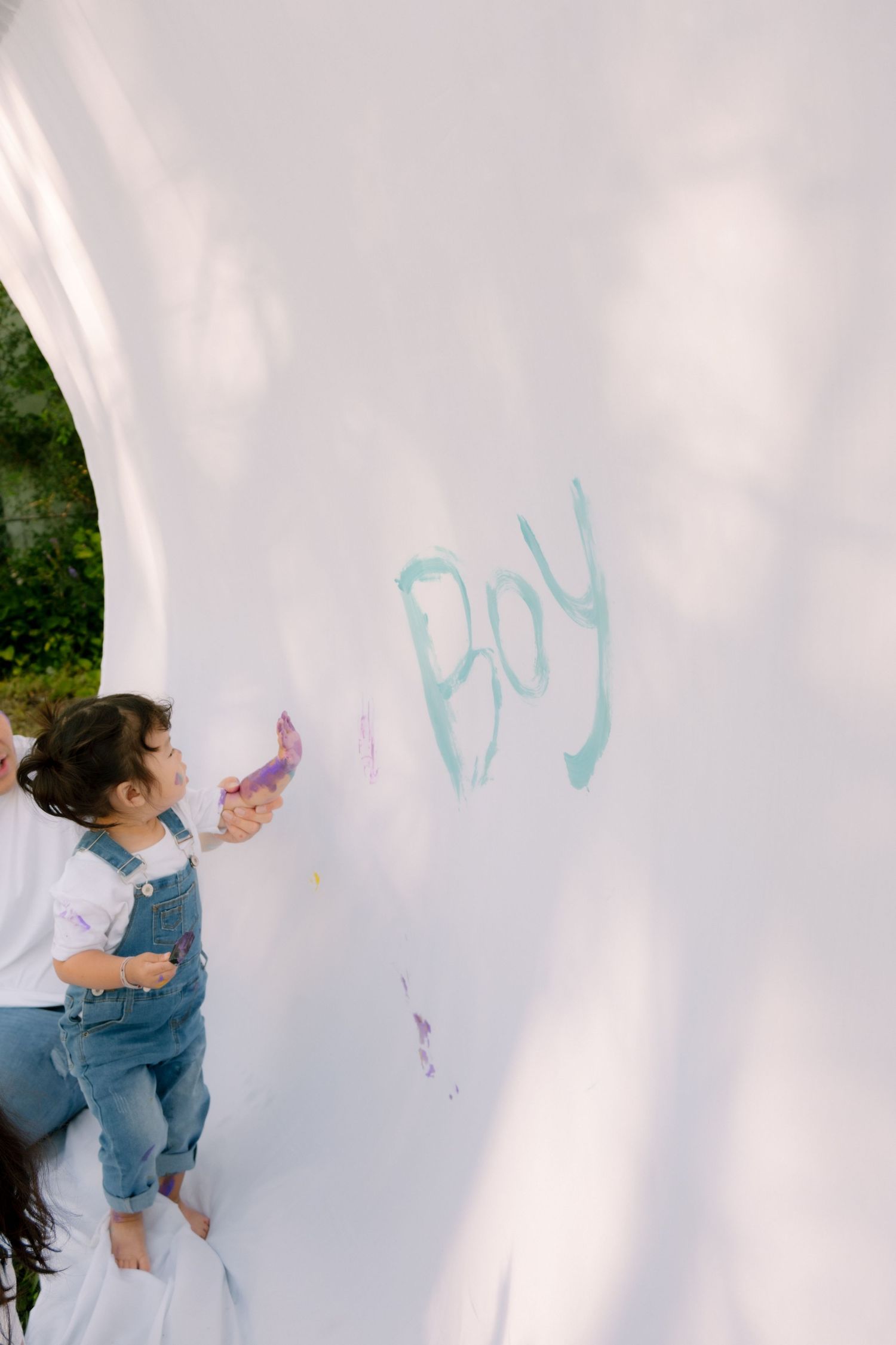 Young child in denim overalls painting the word Boy on large white canvas outdoors.