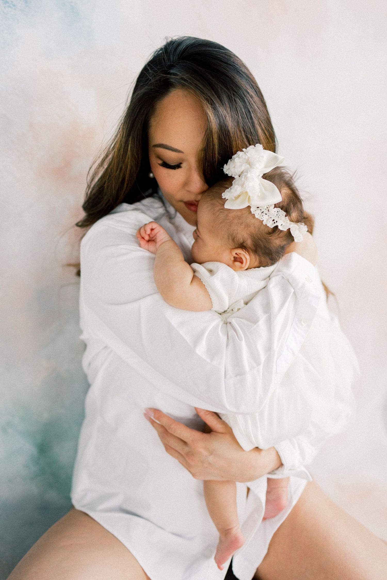 A mother in white gently kisses her newborn baby girl wearing a white bow headband against a soft pastel background.