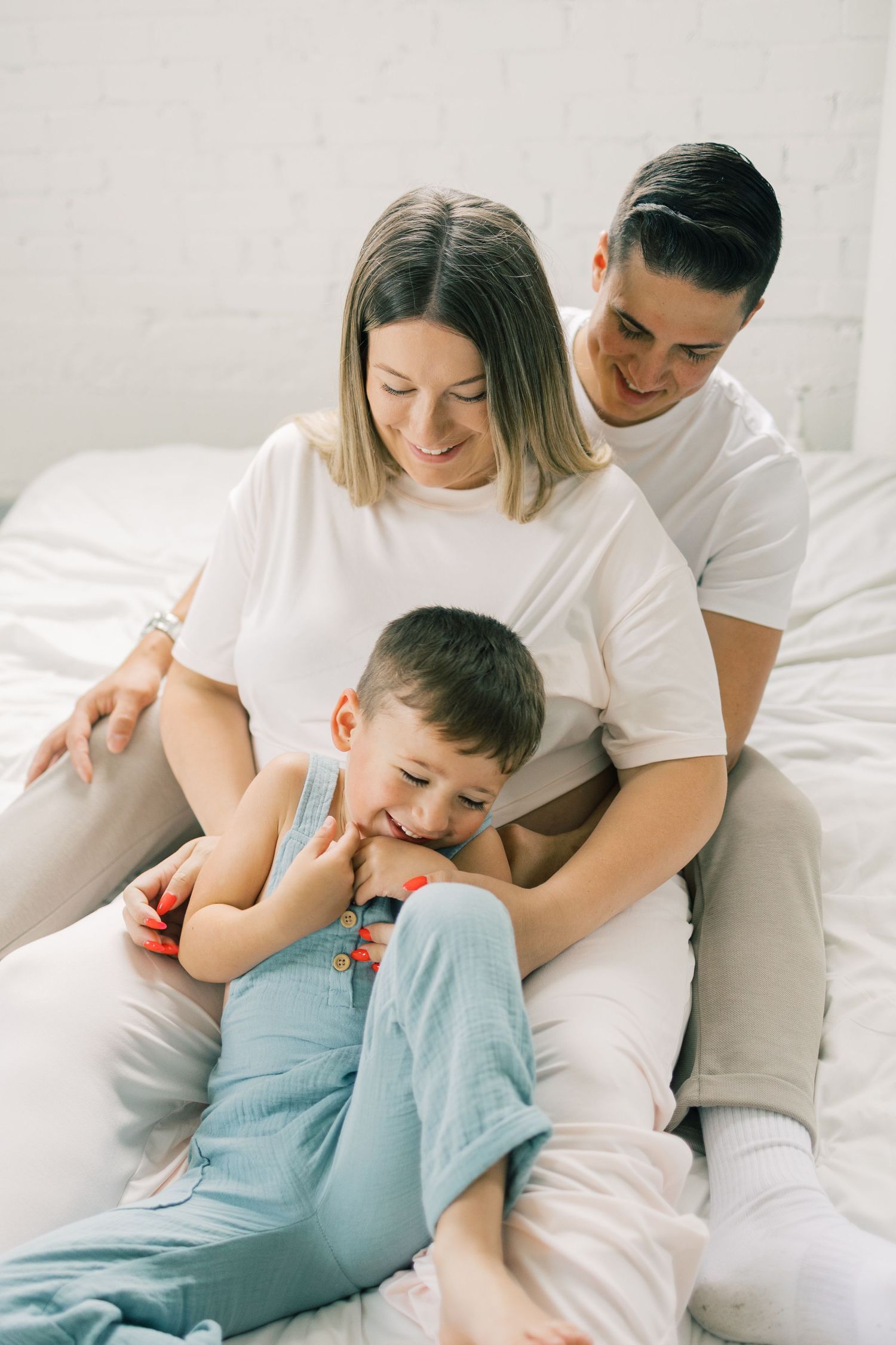 Happy family with two parents and young child sitting together on a white bed, smiling and sharing a joyful moment.