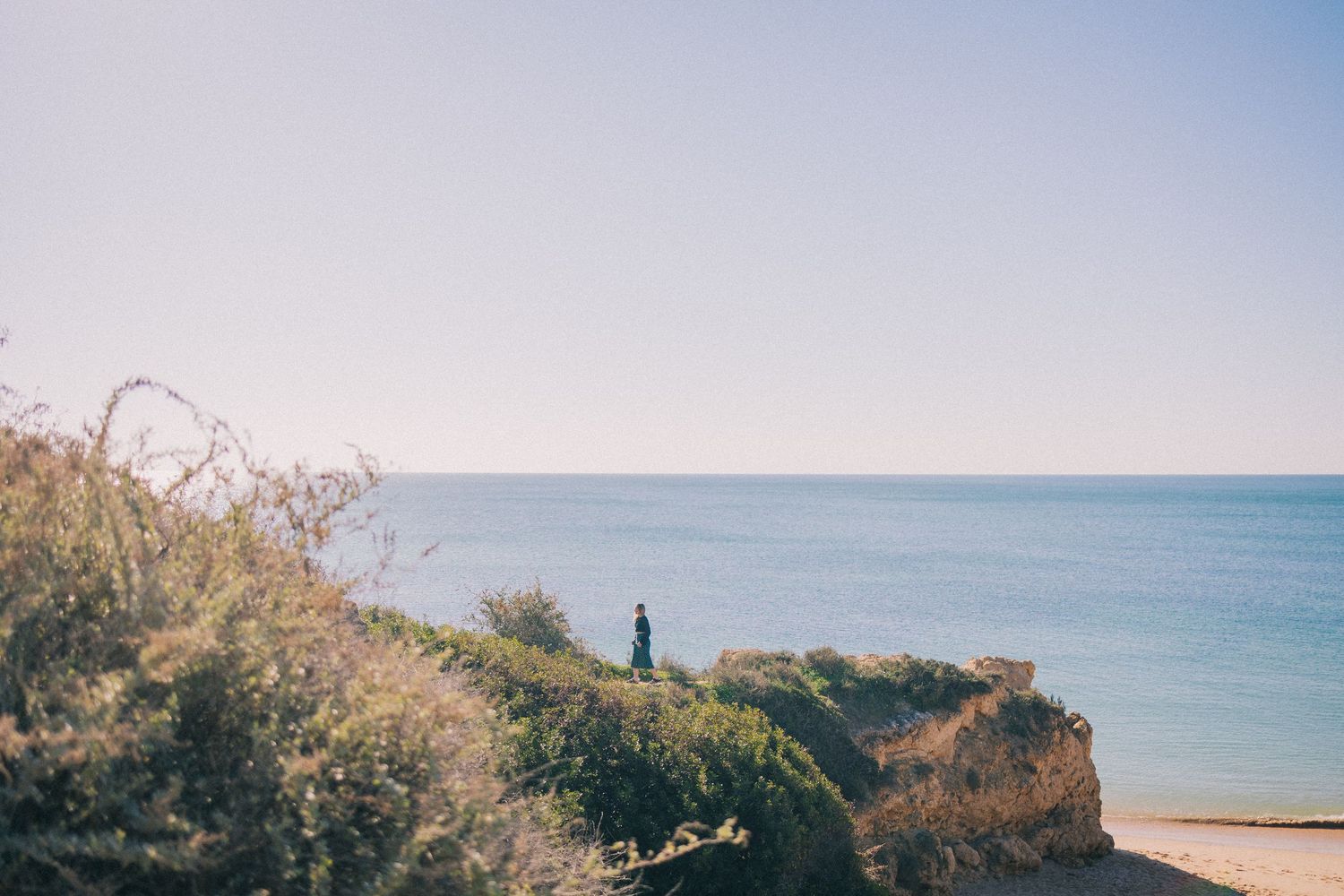 A lone figure stands on a coastal cliff overlooking a calm blue sea, surrounded by lush green shrubs under a clear sky.