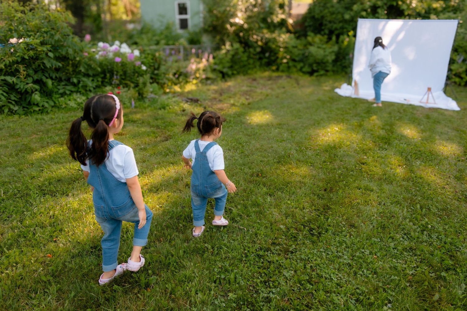 Two young girls in matching denim overalls walk toward a white backdrop in a lush green garden.