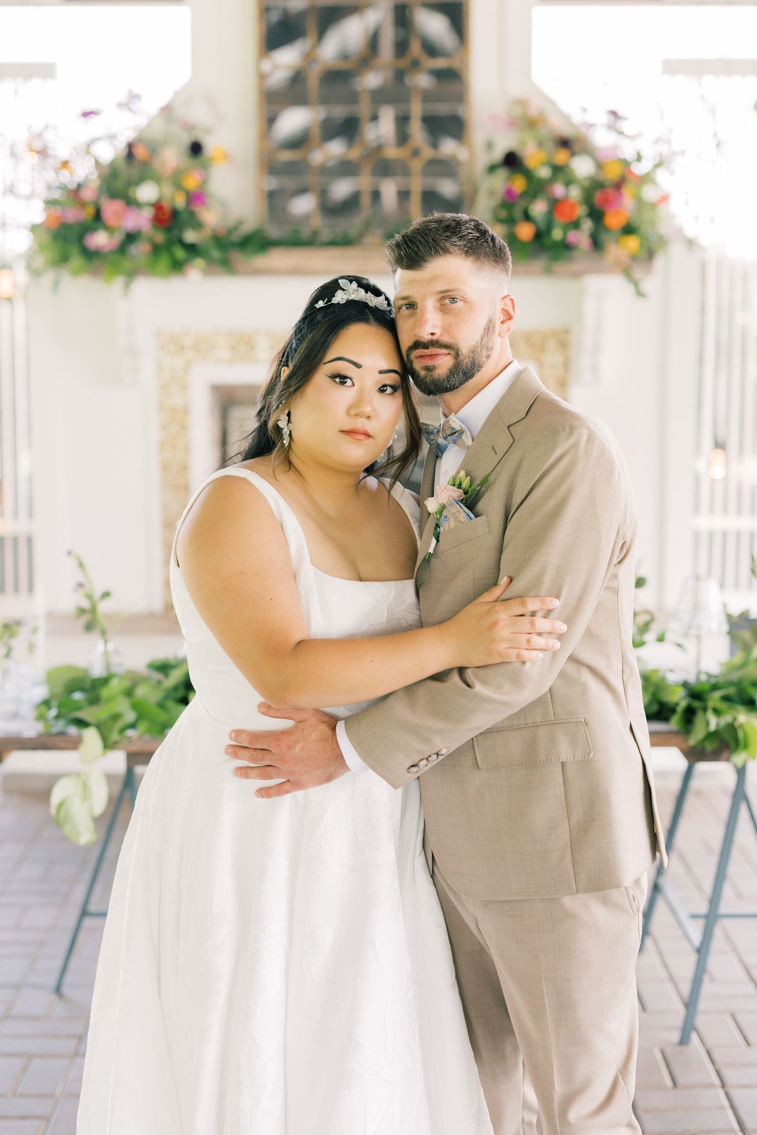A romantic wedding portrait of a couple in white dress and tan suit against a floral backdrop.