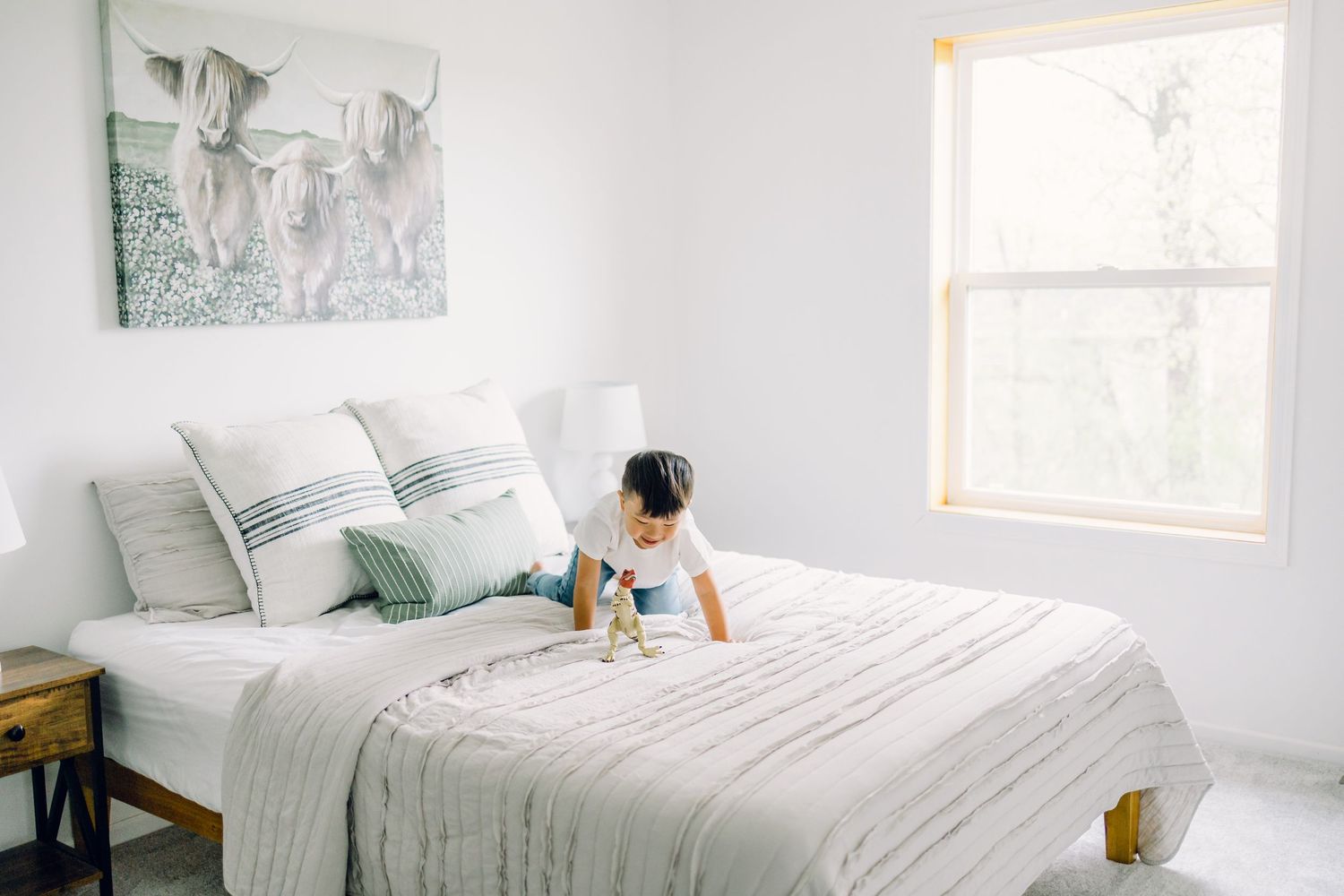 A bright white bedroom with a cozy bed, window, and teal accents features someone sitting on fresh linens.