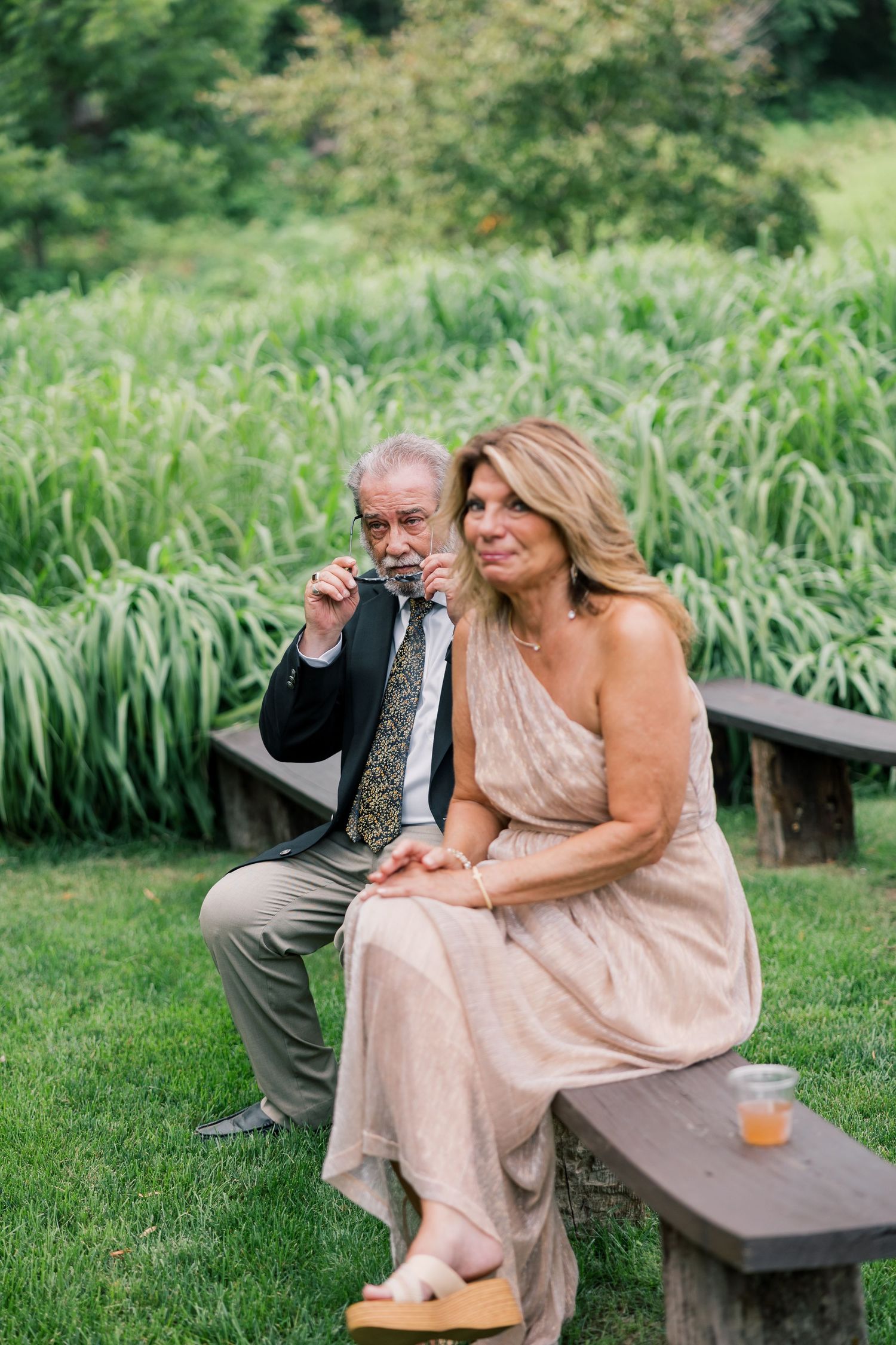 Wedding guests relax on wooden benches in formal attire during outdoor reception.