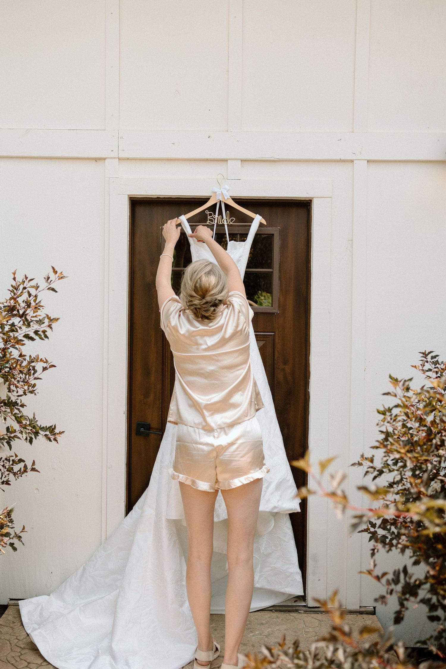 White wedding dress hanging outdoors against a light-colored wall with foliage.