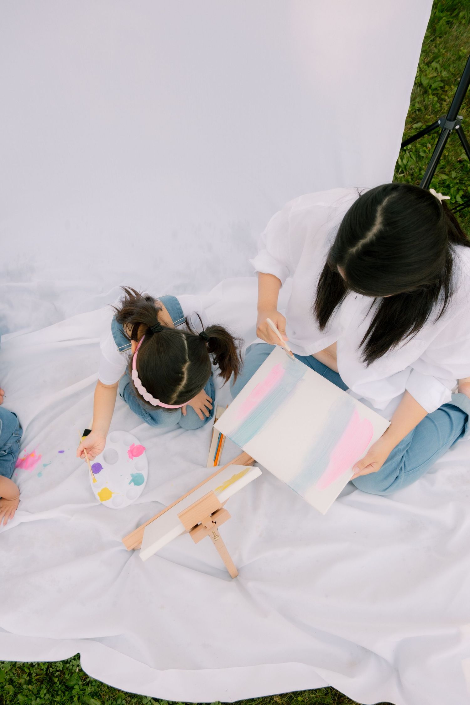 Mother and daughter painting together on white blanket outdoors, with colorful palette and canvas on small easel.