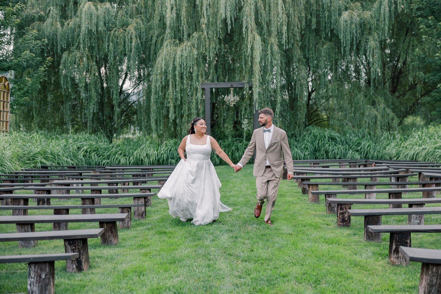 Wooden benches line a garden ceremony space under weeping willow trees creating a romantic outdoor setting.