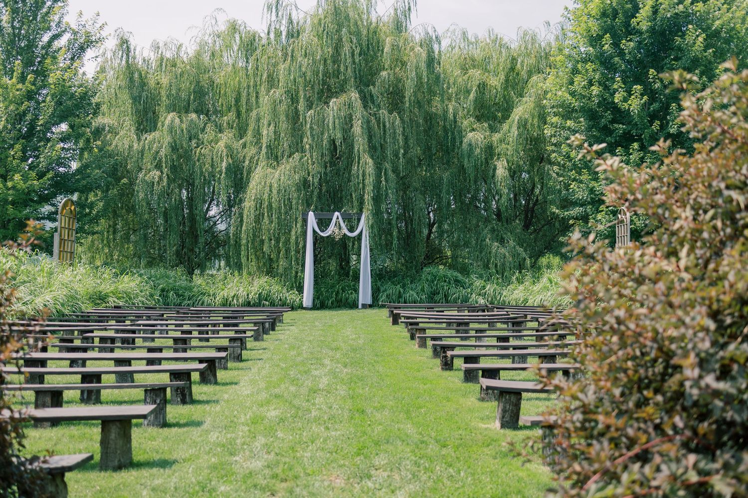Outdoor wedding ceremony site with wooden benches arranged on a green lawn surrounded by trees.