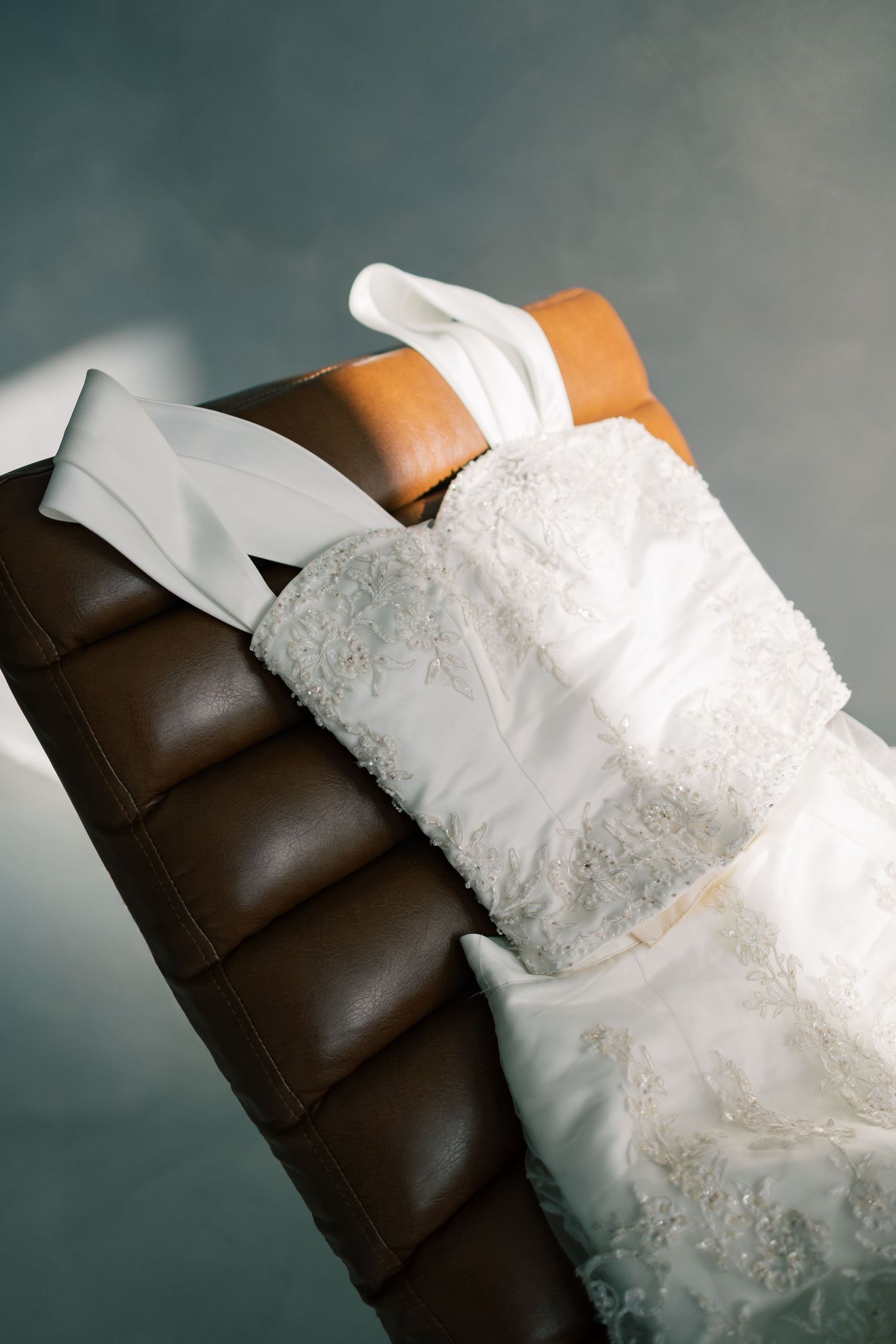 A delicate white lace wedding dress draped over a dark brown leather couch in a studio setting.