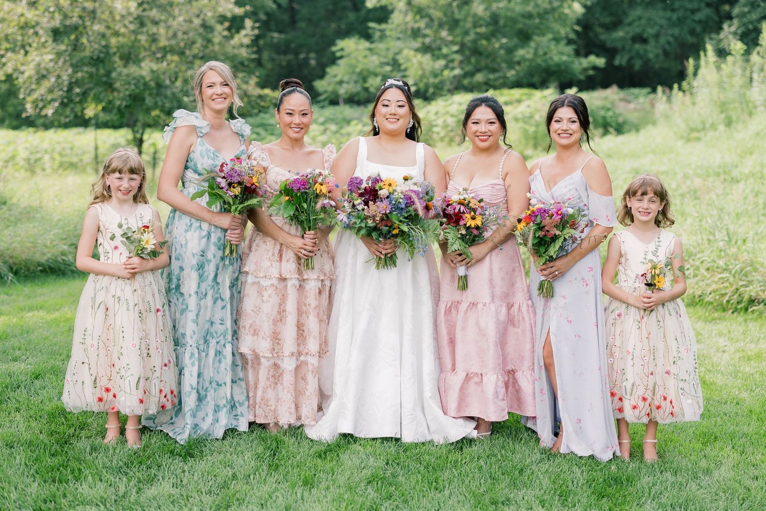 Bridesmaids in pastel floral dresses pose together on a lush green hillside.