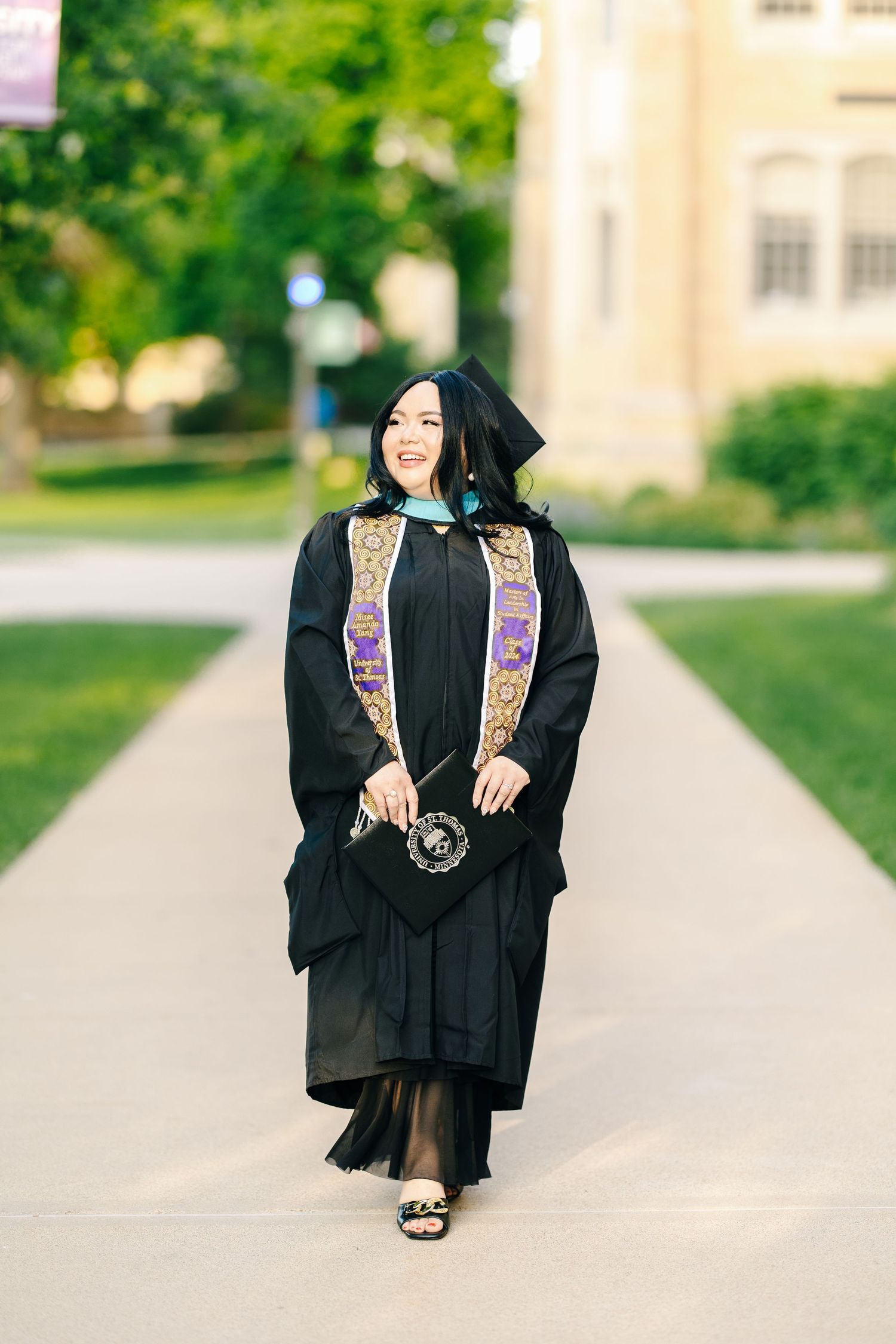 A graduate in black cap and gown with decorative stoles walks along a campus pathway on a sunny day.