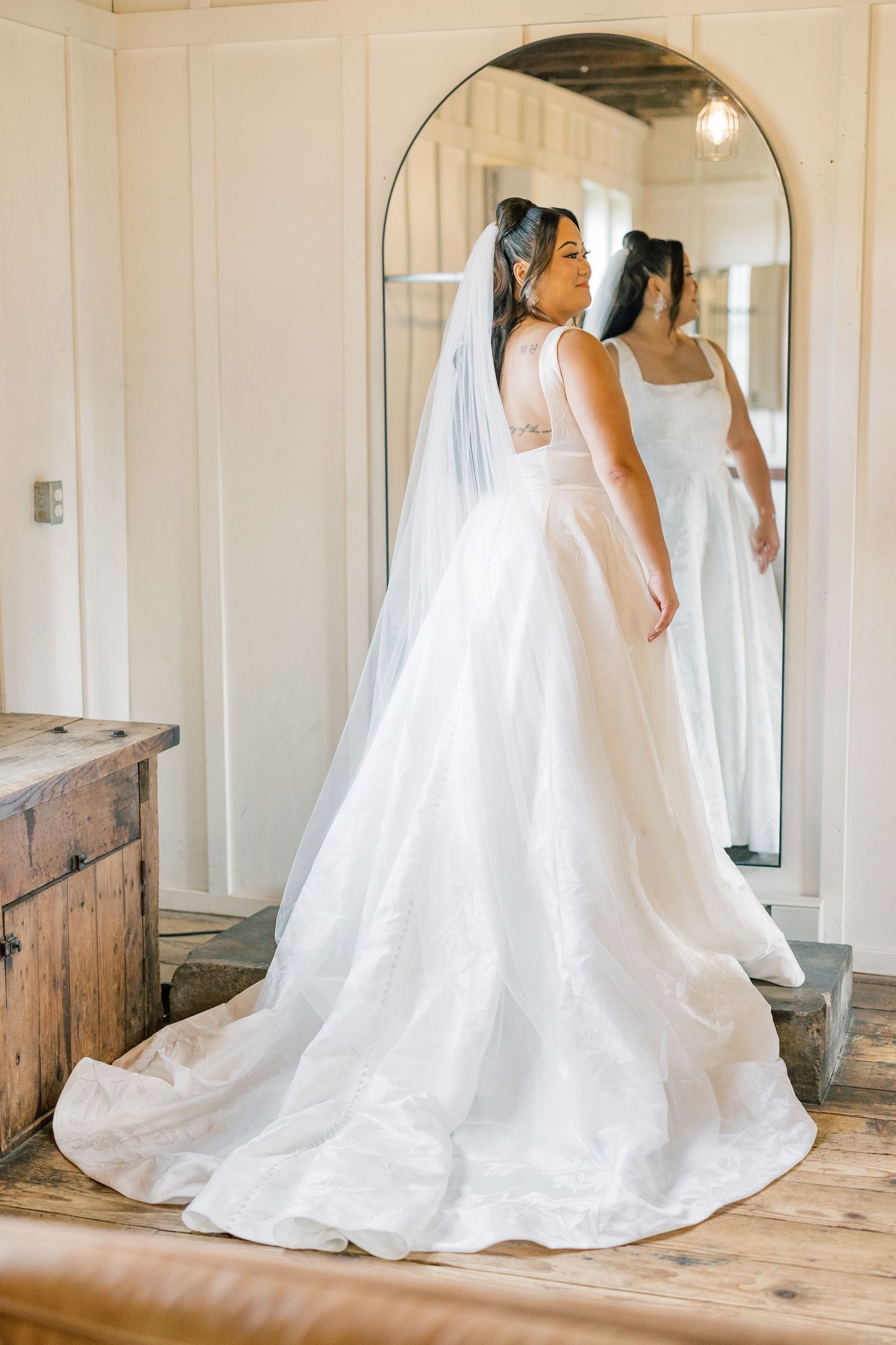 A bride in a flowing white wedding gown looks at her reflection in an ornate mirror.