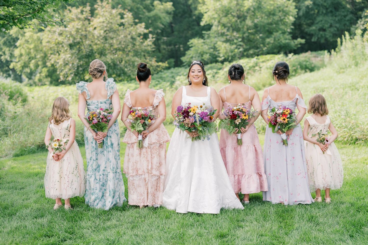 Bridesmaids in pastel floral dresses stand together holding bouquets outdoors.