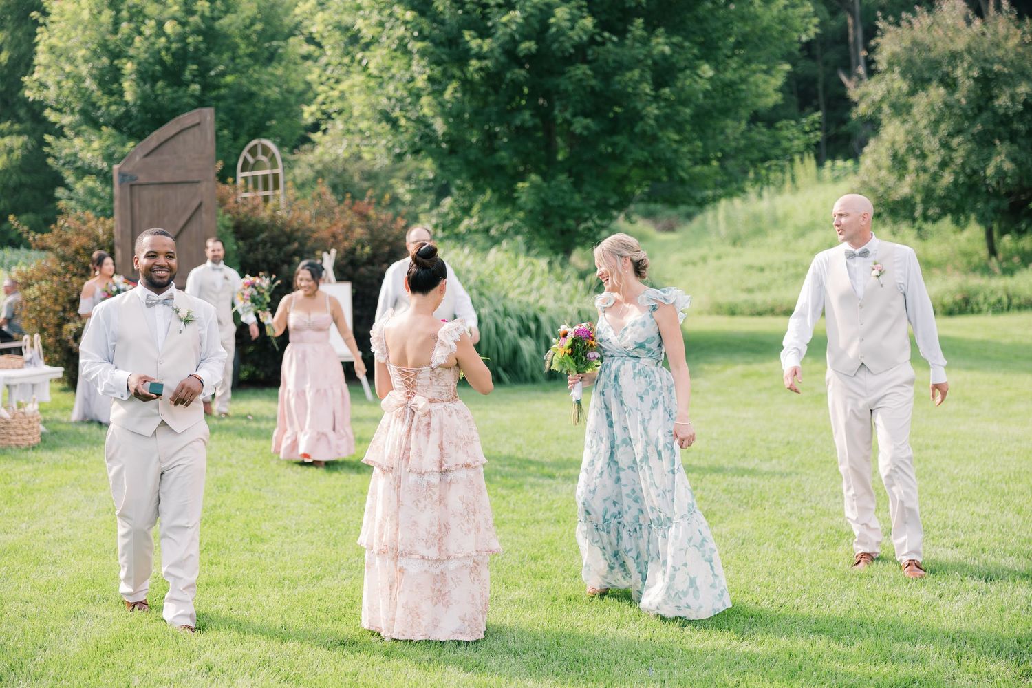 Wedding party members in pastel dresses and light suits walk across a sunny lawn.