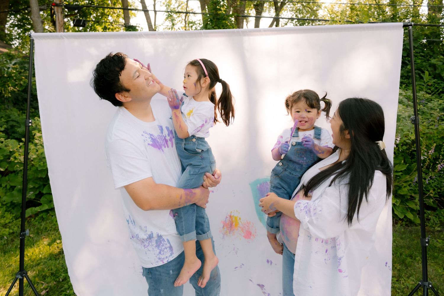Happy family with two young daughters covered in paint laughing together outdoors against white backdrop.