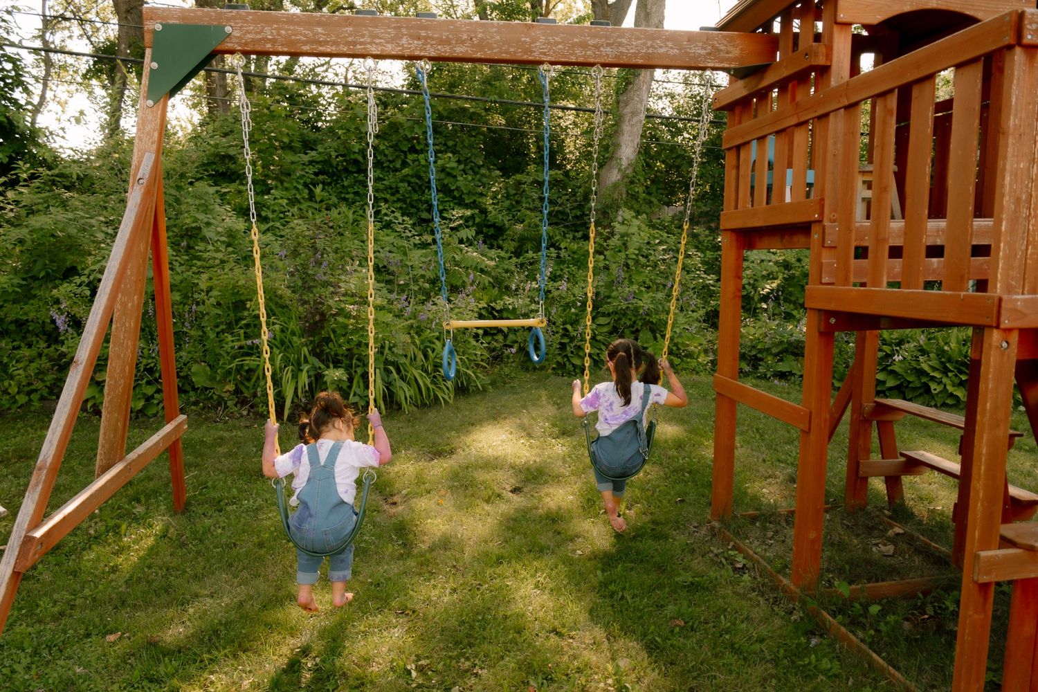 Two young girls swinging on a wooden backyard swing set surrounded by lush green trees and grass.