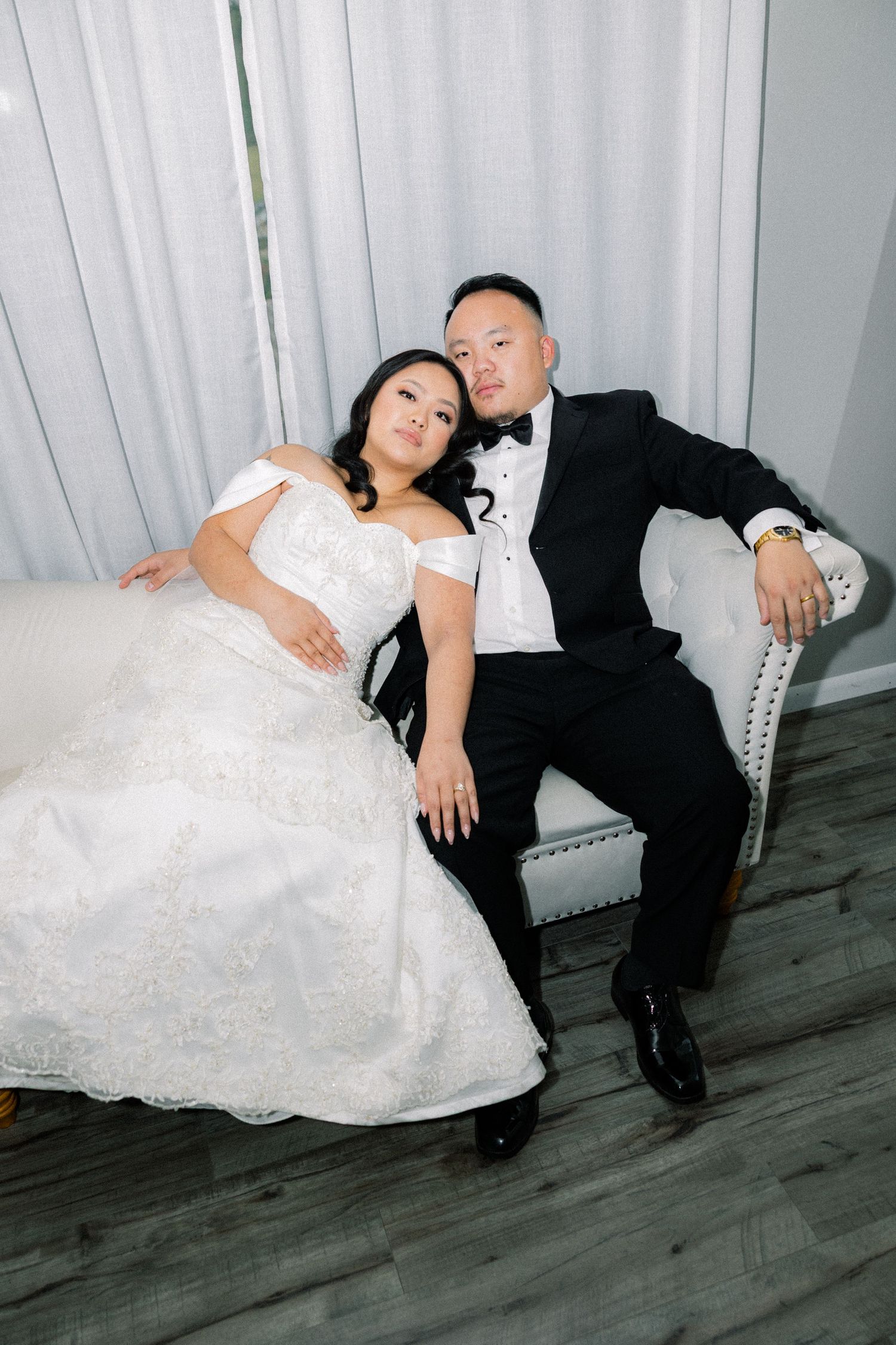 A bride in a white lace gown and groom in a black tuxedo relax together on an elegant gray upholstered chair.