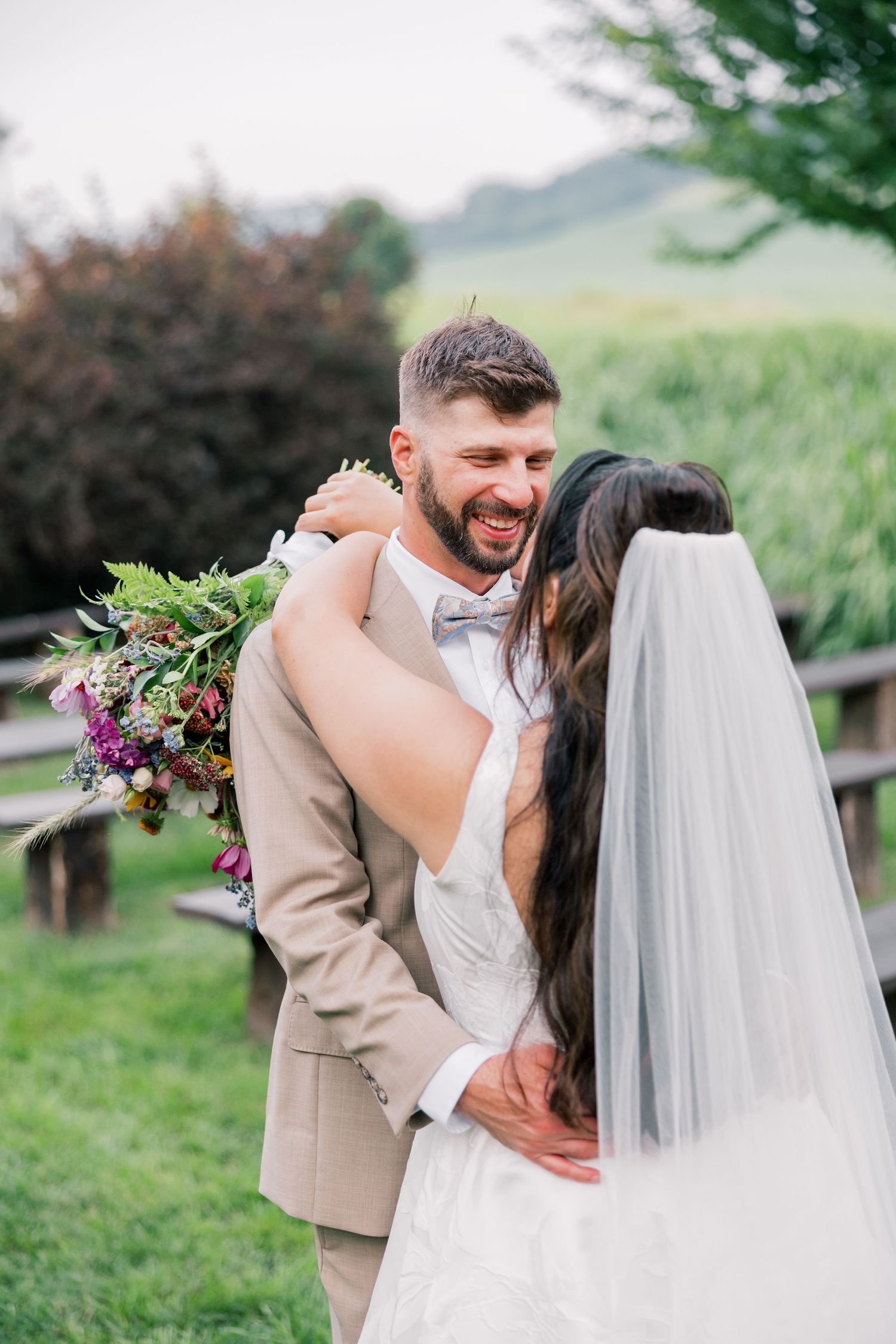 Romantic wedding couple embraces in a garden setting with flowing white veil and tan suit.