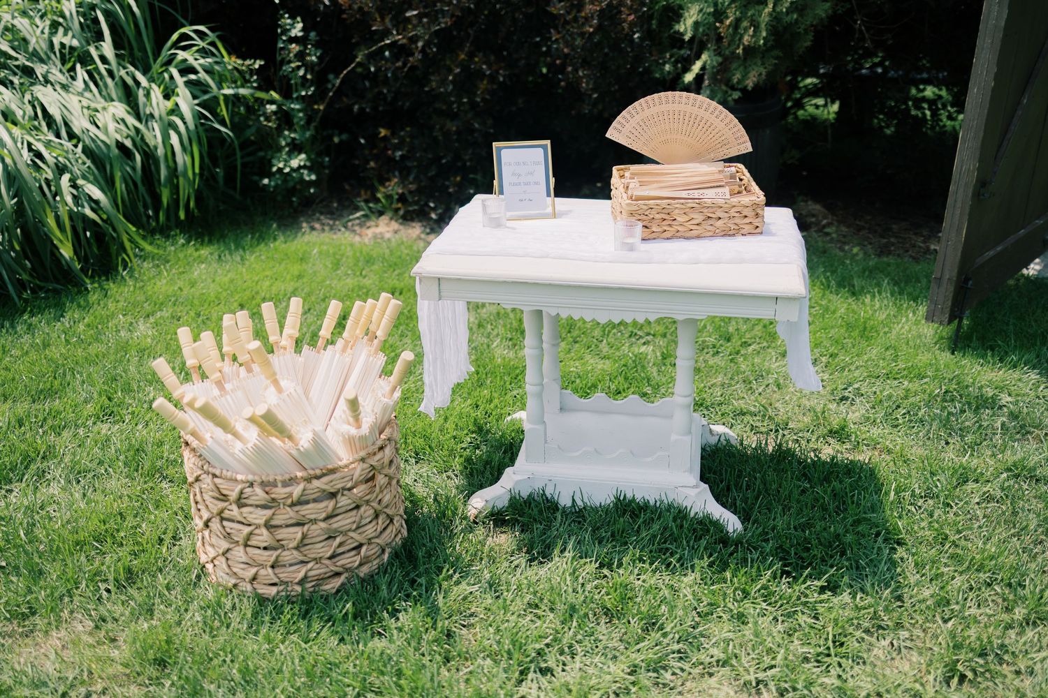 Outdoor wedding reception setup with white furniture and woven baskets on a sunny lawn.
