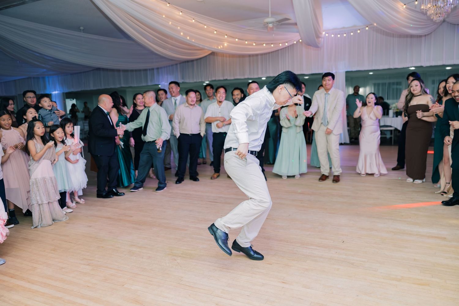 Wedding guests dance and celebrate on a ballroom floor during a reception party with white draped decor.