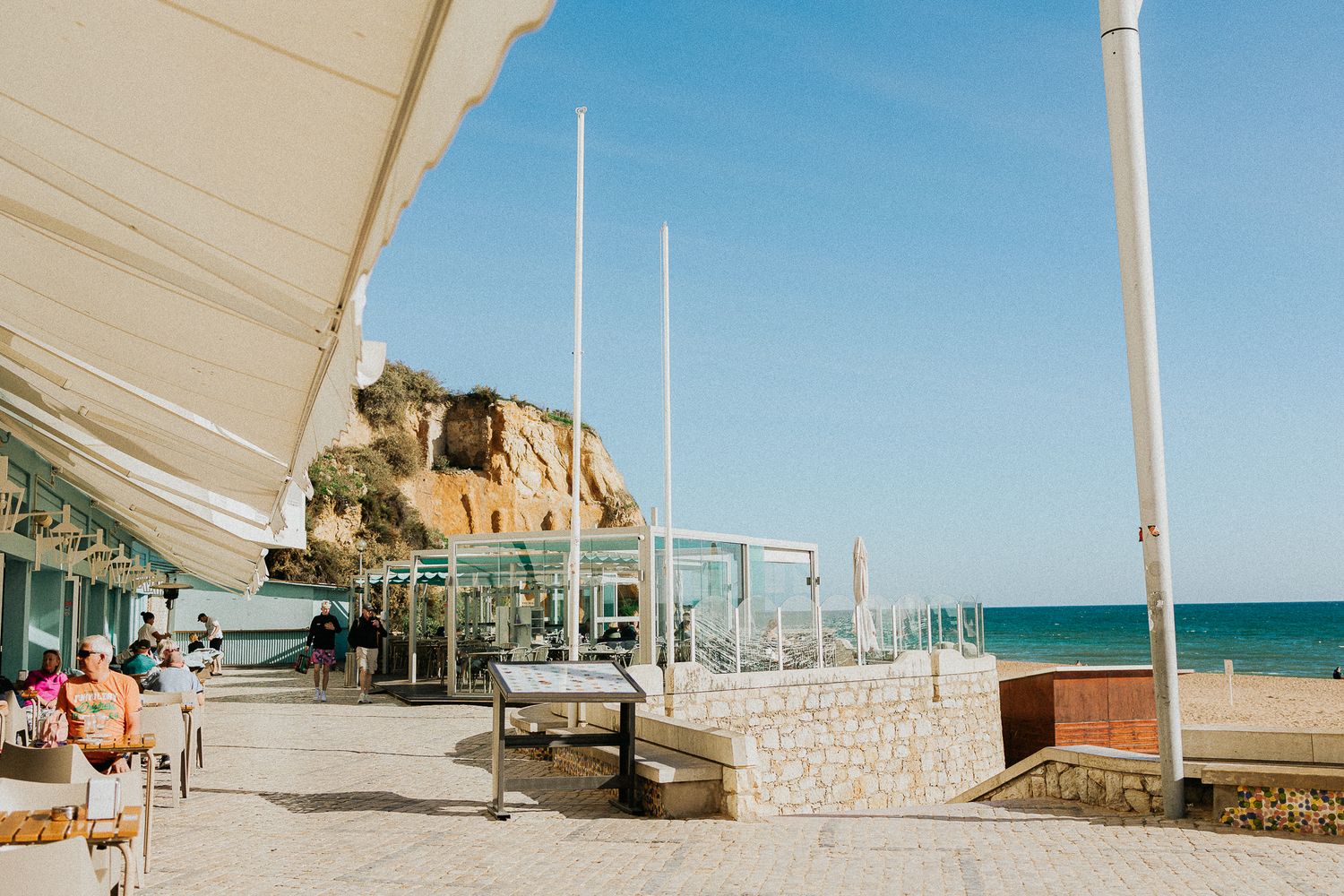 Beachfront promenade with open-air restaurant, stone walls, and blue sea views on a sunny day.