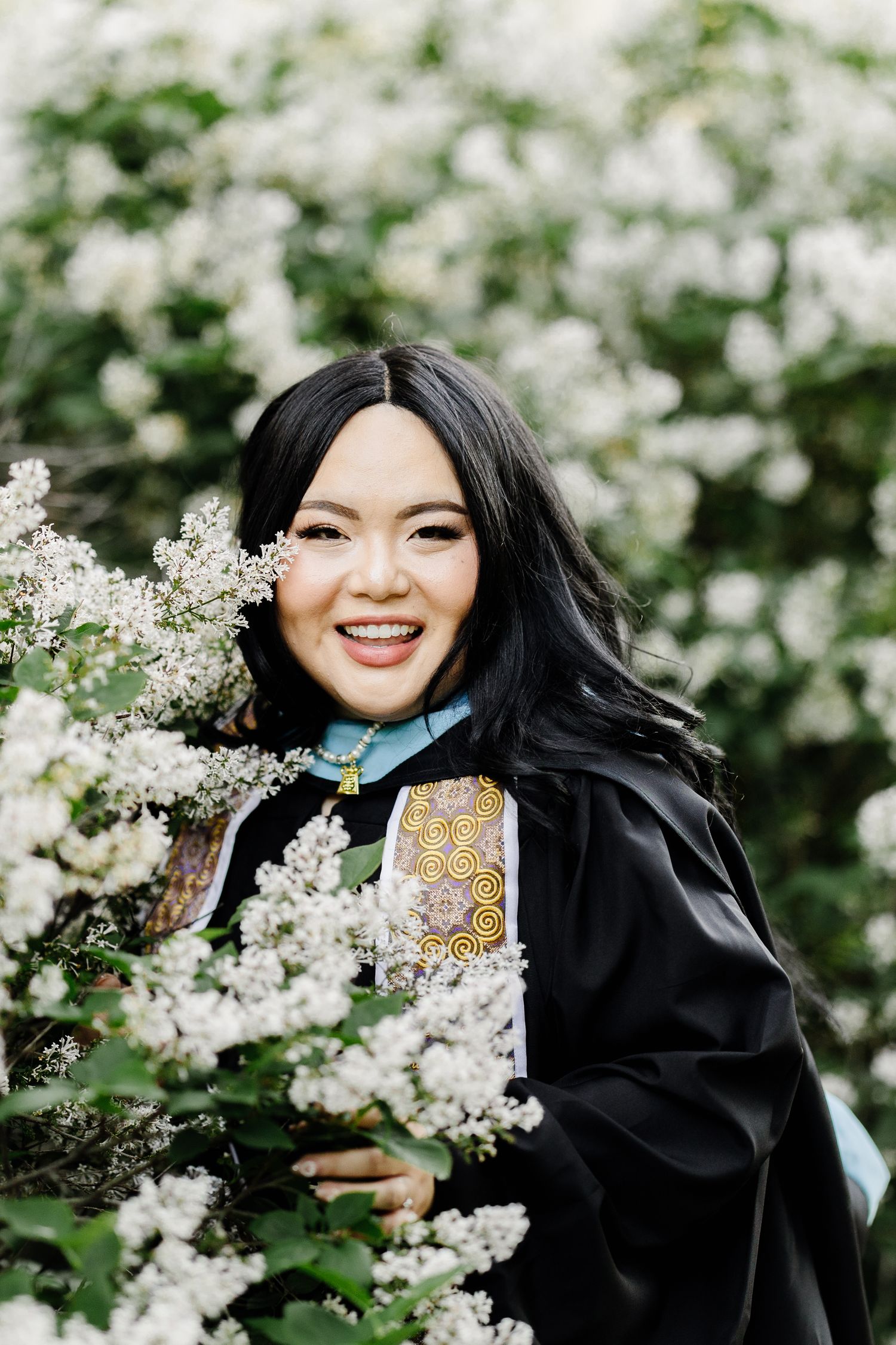 Graduate in academic regalia poses beside blooming white flowers in spring.