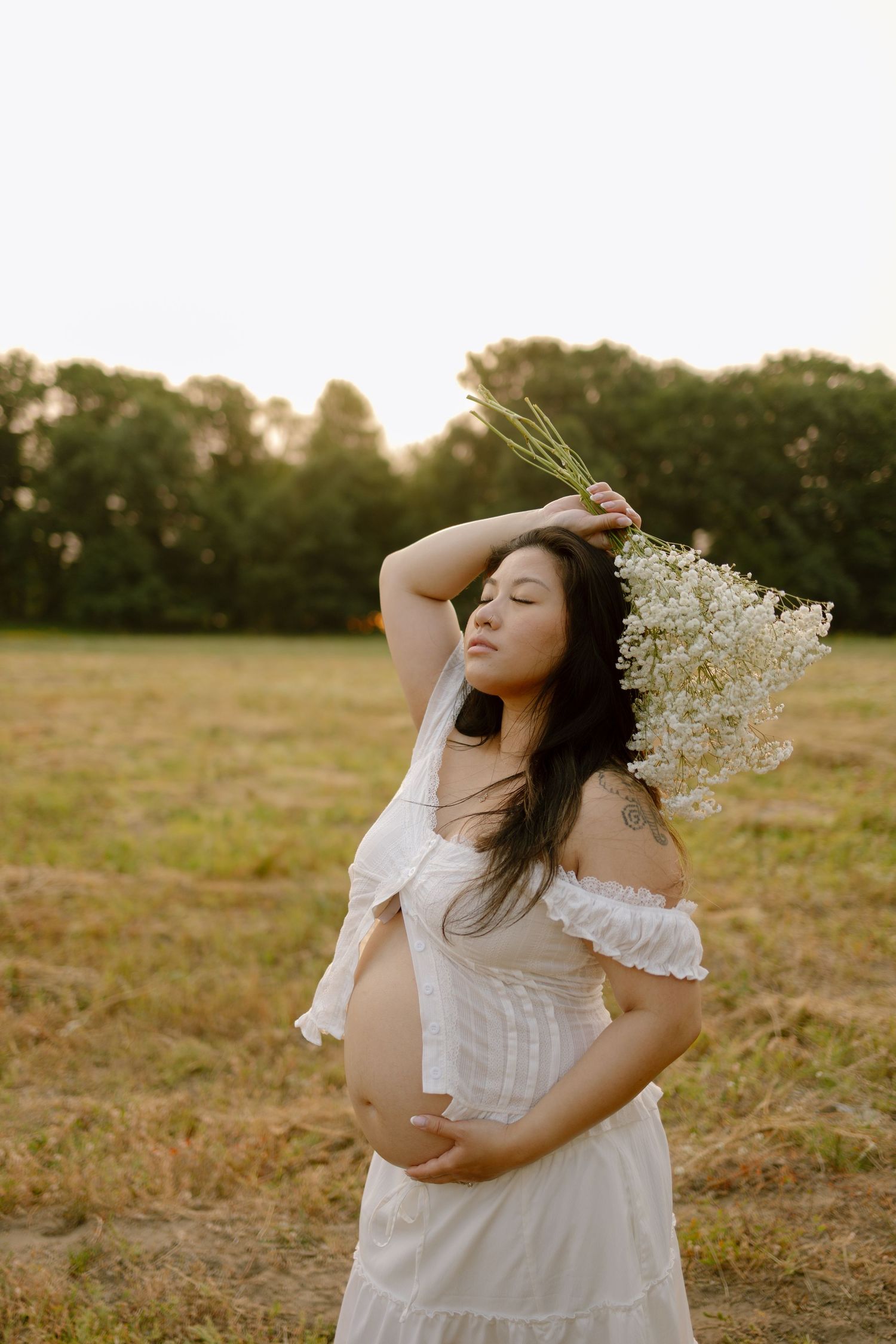 Pregnant woman in white off-shoulder dress holding wildflowers in a sunlit field at golden hour.