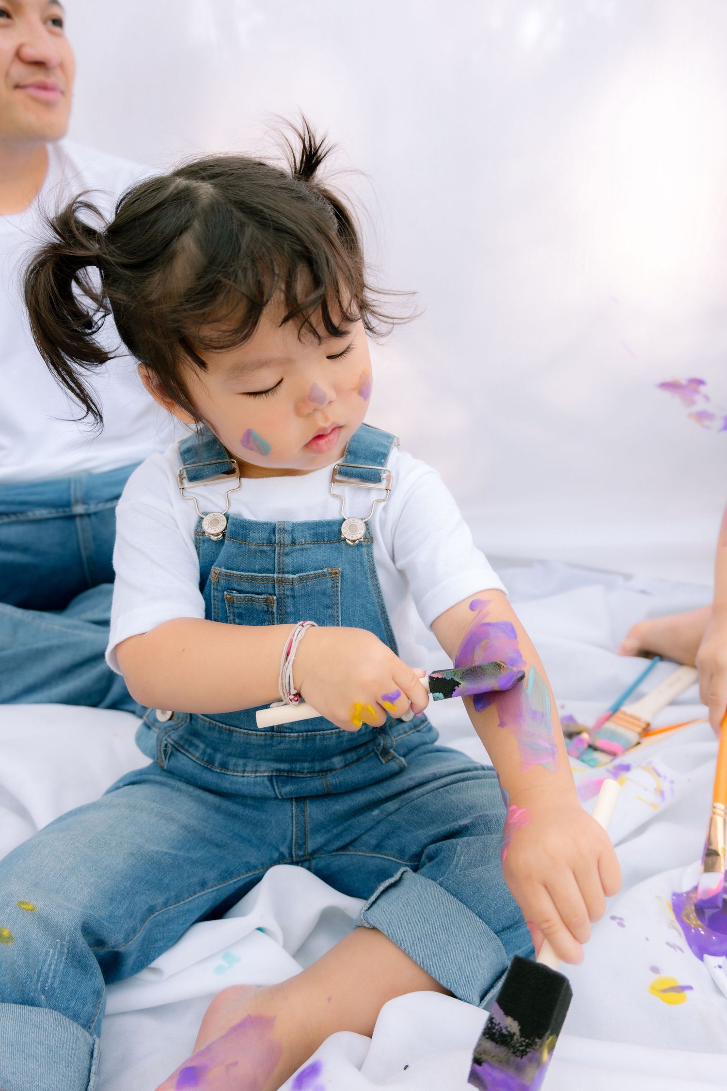Toddler girl in denim overalls covered in colorful paint during a fun outdoor activity.
