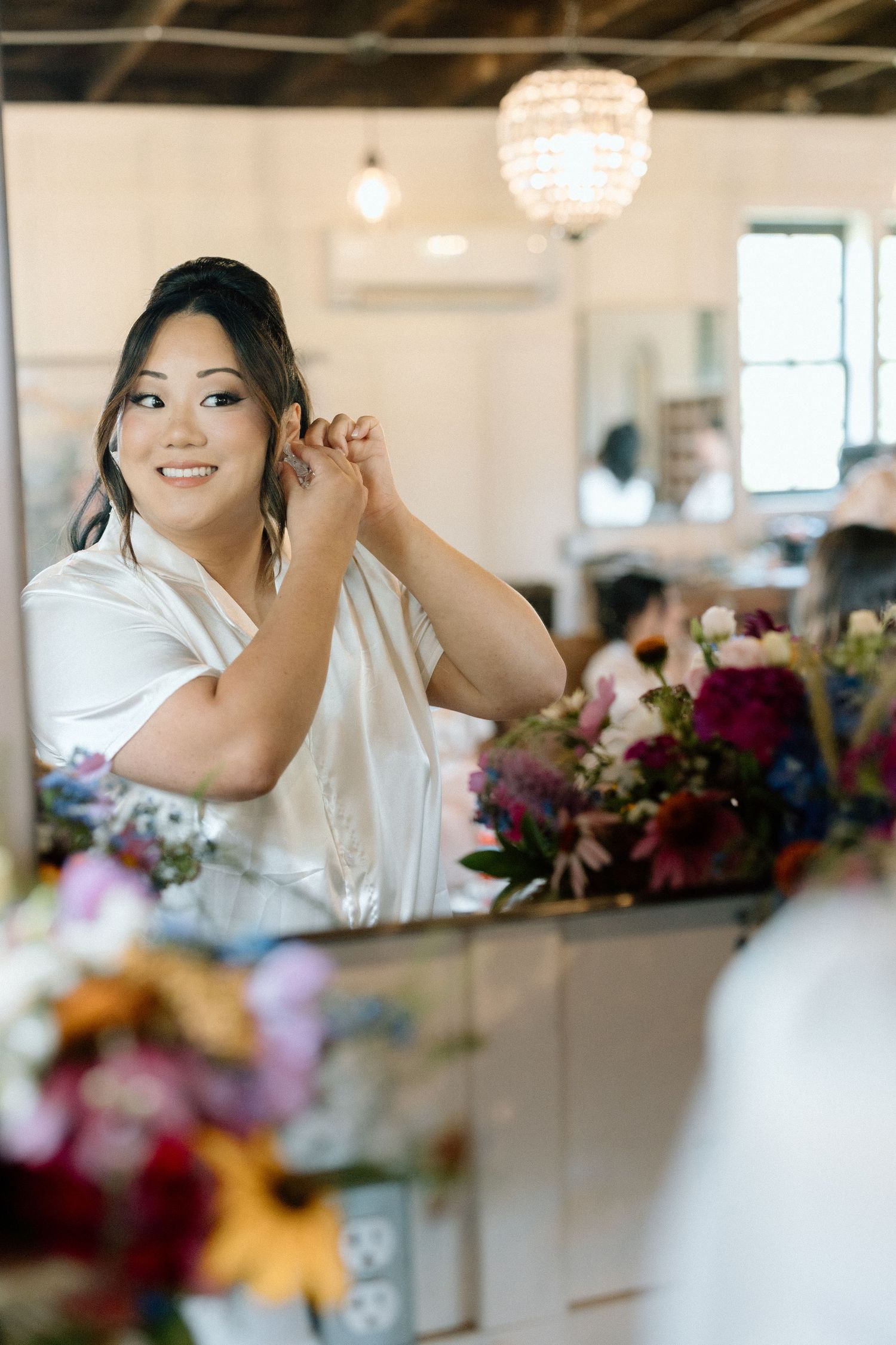 A person puts on earrings while standing near colorful floral arrangements in an indoor venue.