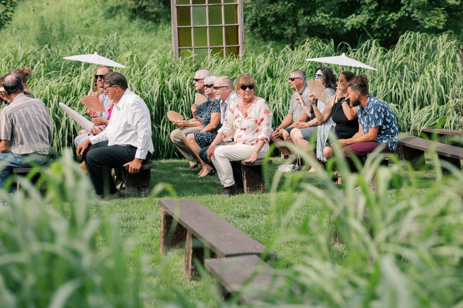Wedding guests relaxing on outdoor benches surrounded by tall grass and greenery.