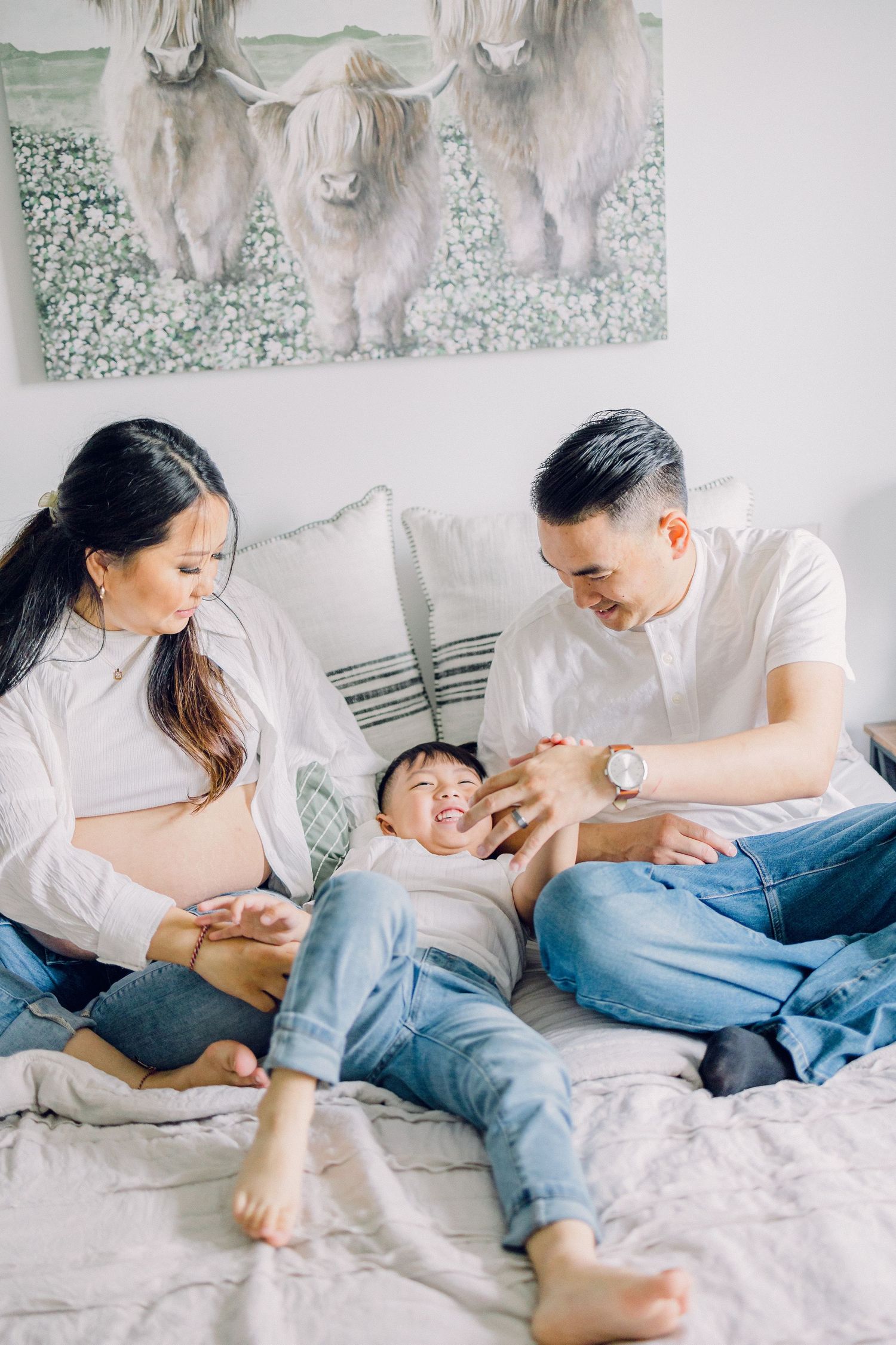 A joyful family moment shared on a bed with a highland cow artwork on the wall in the background.