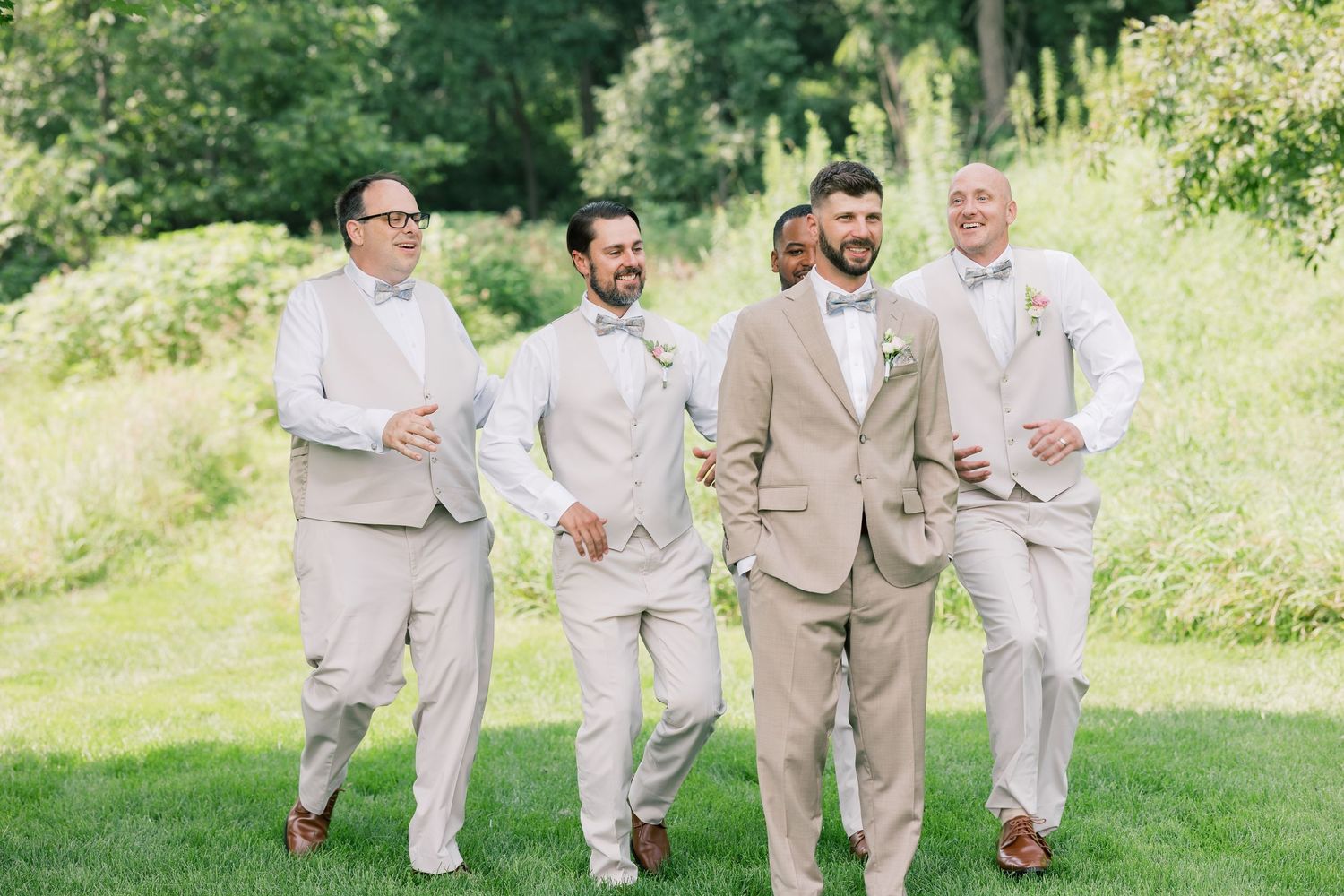 Wedding party in beige and white attire poses together on lush green lawn.