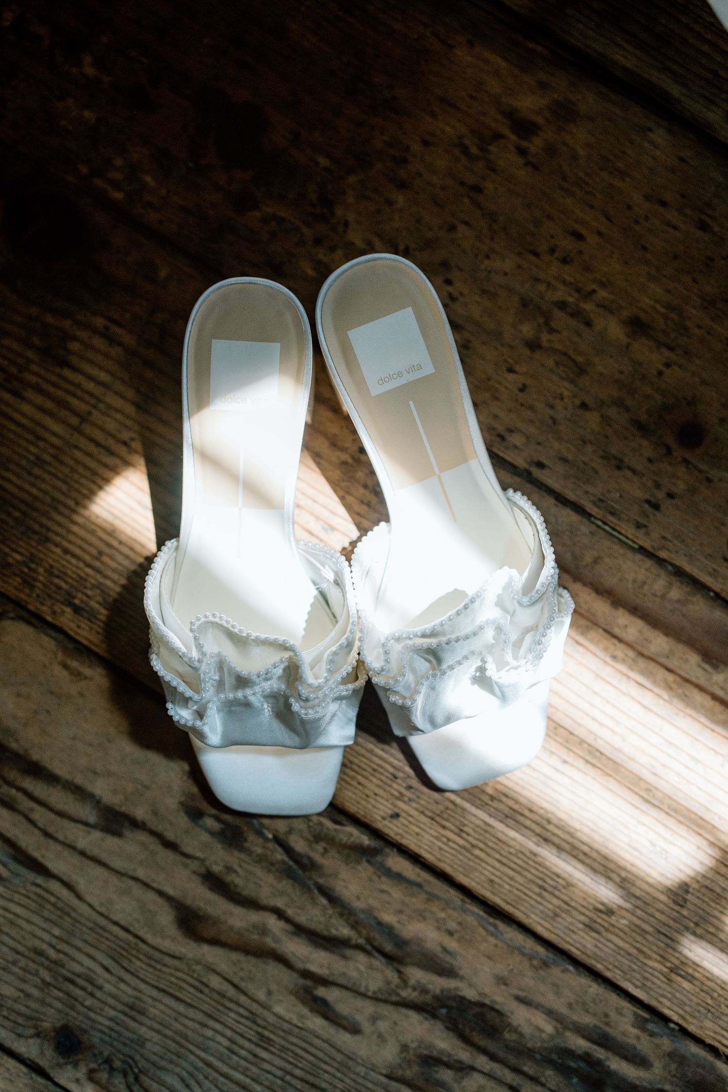 White bridal shoes with ruffled details sit on rustic wooden floorboards in natural lighting.