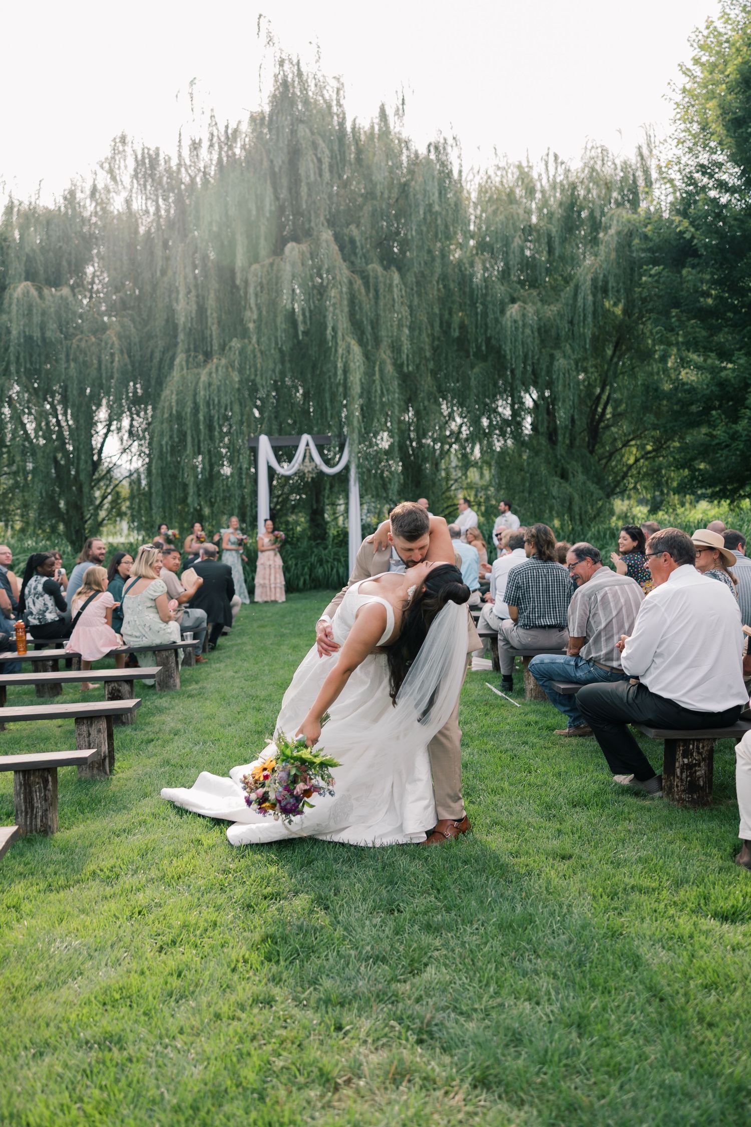 Couple shares romantic dance moves on grass with wedding guests watching from wooden benches.