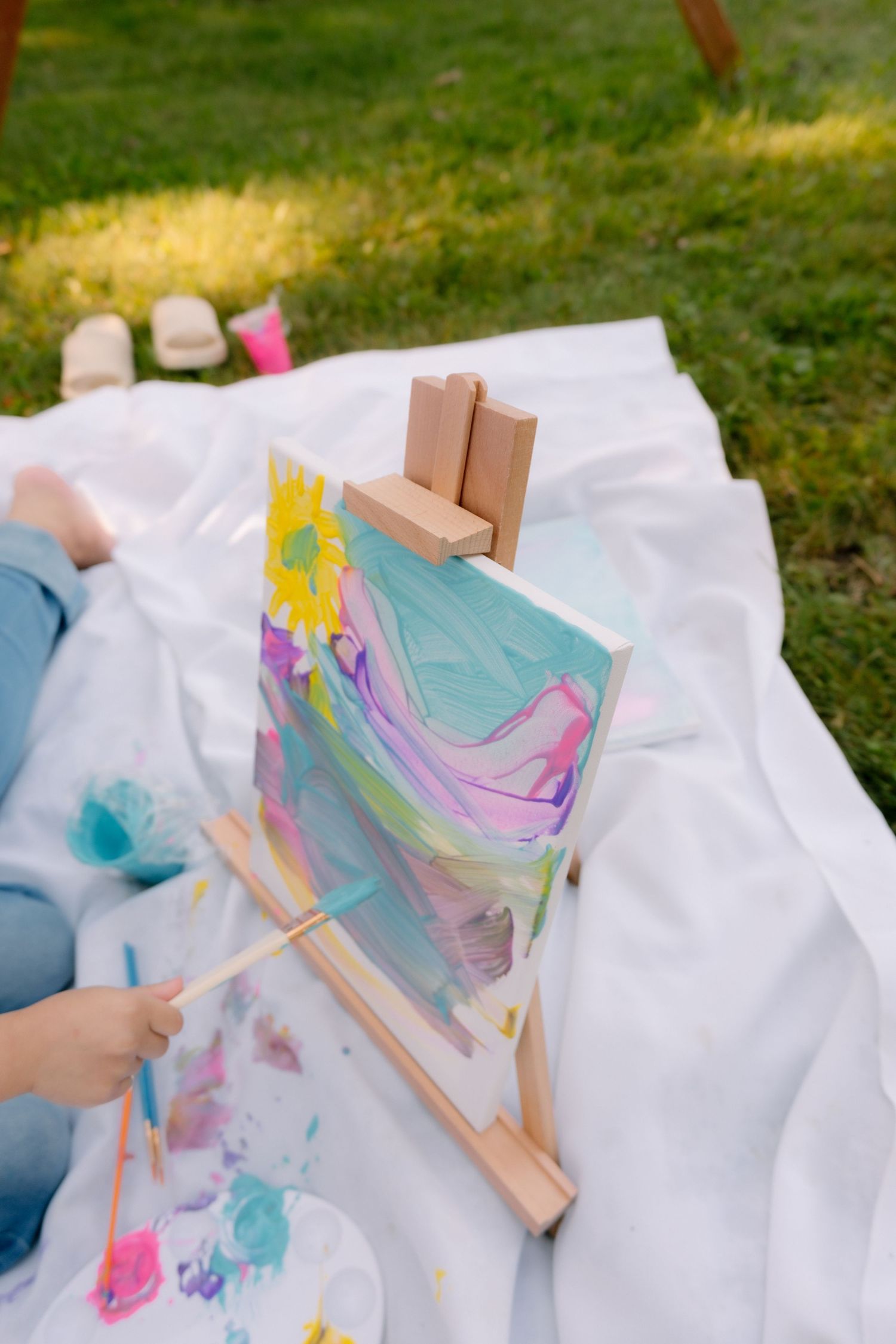 Child painting colorful abstract art on small easel outdoors on white blanket in garden.