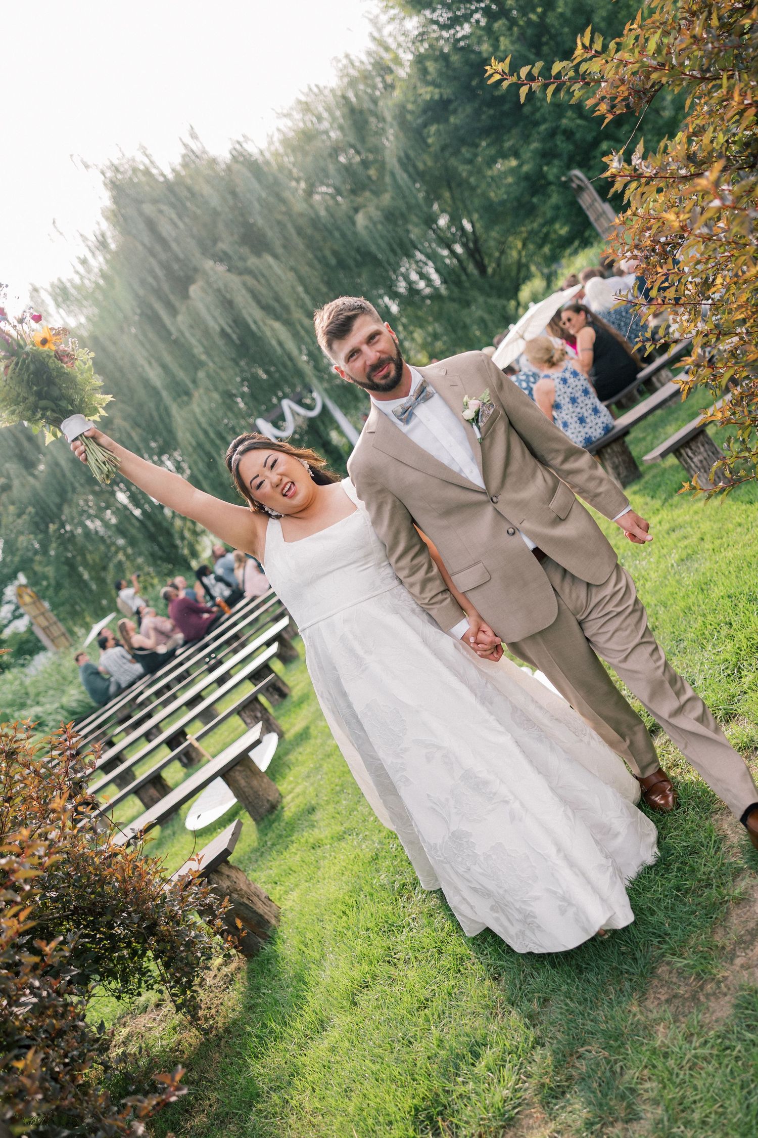 A couple in wedding attire dances and celebrates joyfully on a grassy lawn with autumn trees in the background.
