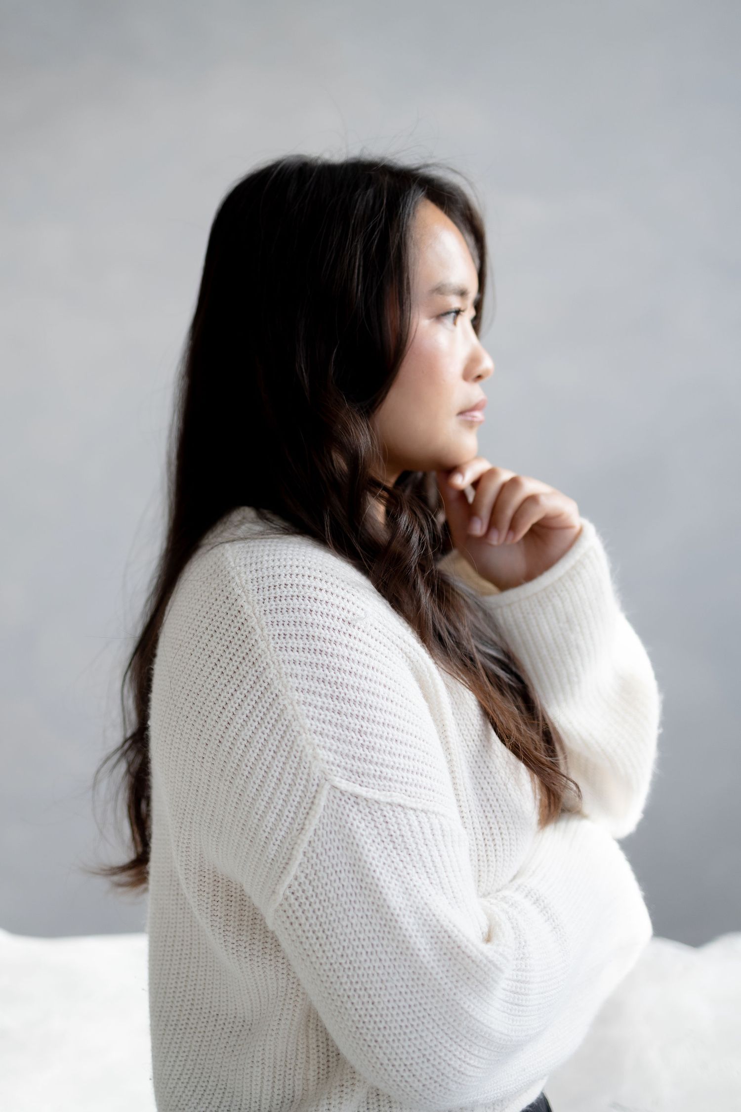 A thoughtful young Asian woman with long dark hair in a white knit sweater gazes sideways with hand near chin.