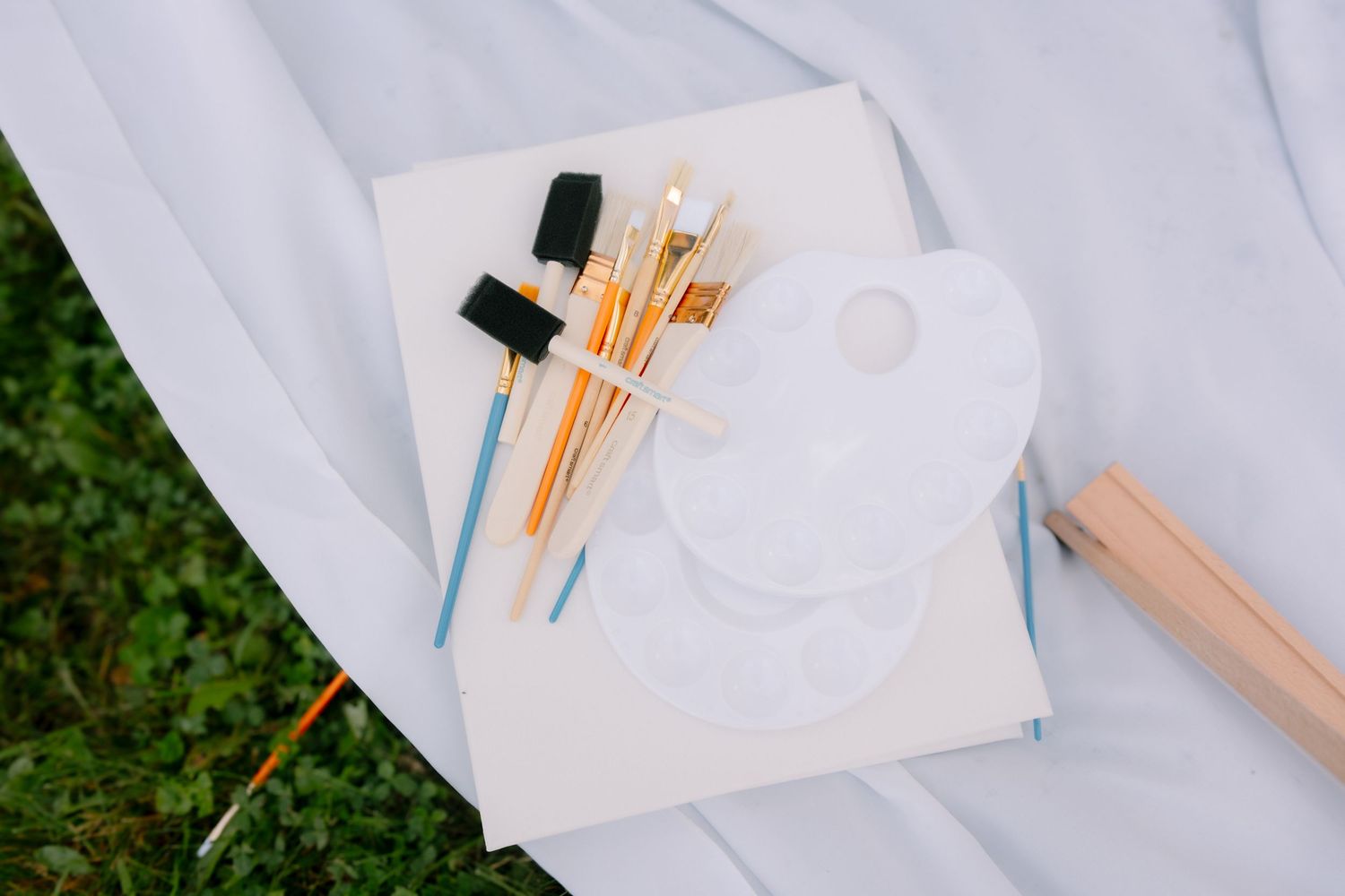 White palette and assorted paint brushes resting on a sketchbook atop a white cloth outdoors.
