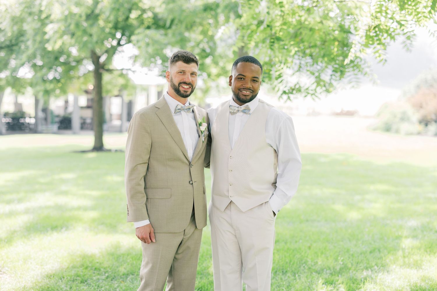 Two people in coordinating beige and white suits stand together in a sunlit outdoor garden setting.