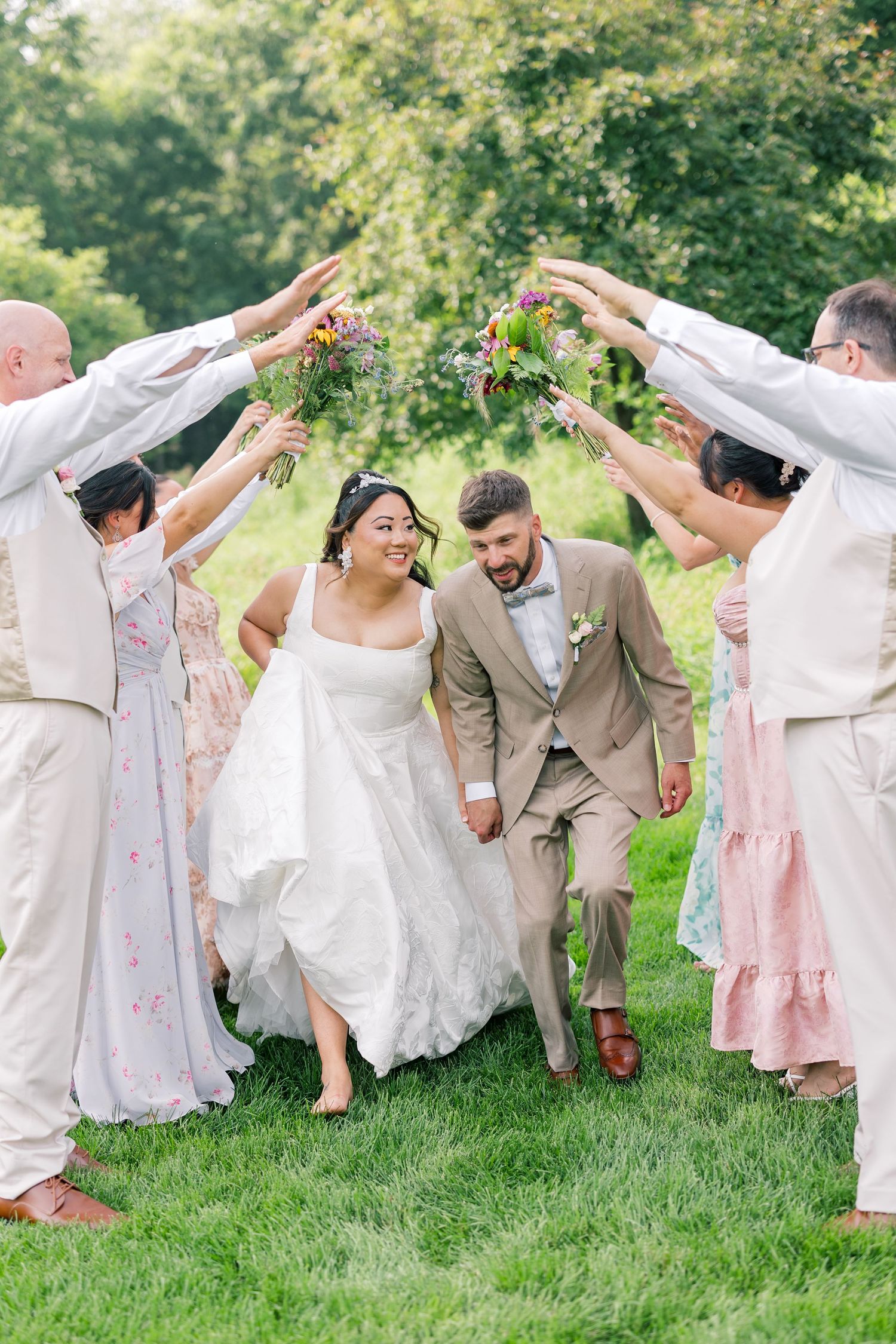 Wedding party in pink and beige attire poses playfully on green grass with raised arms.