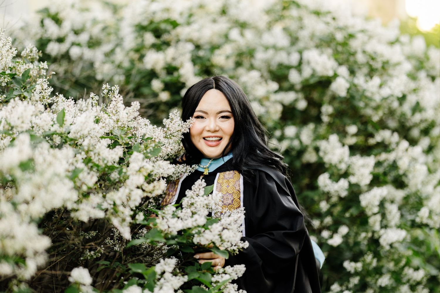 Graduate in black academic regalia poses with white flowering bushes during graduation photo session.