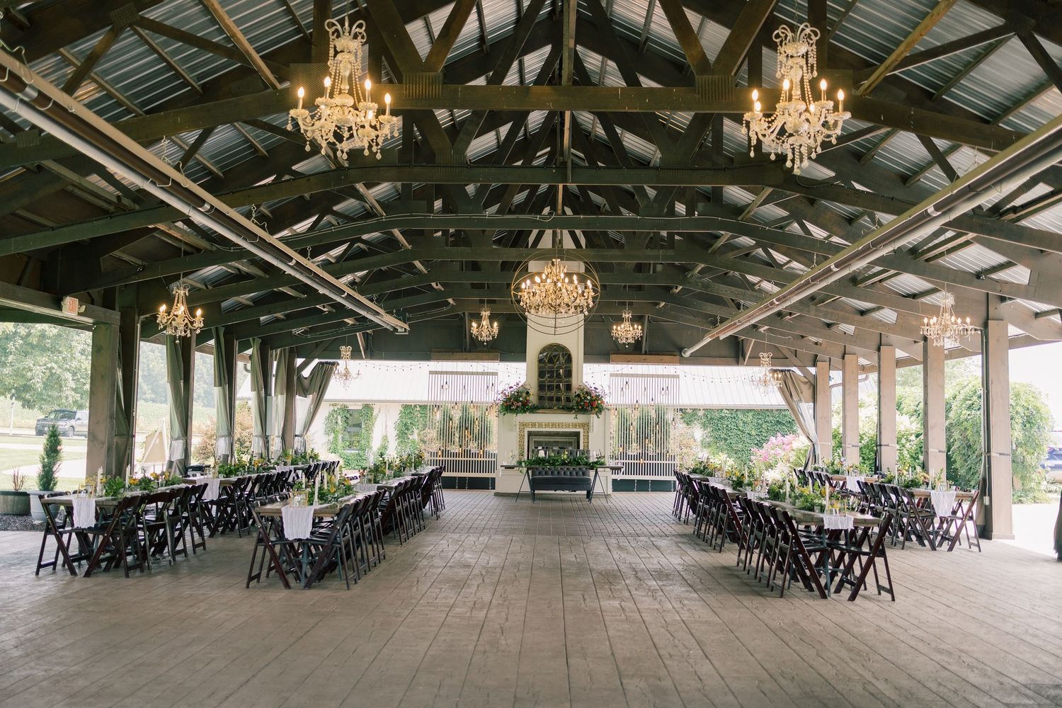 Long rustic reception venue with wooden beams and elegant chandeliers displays rows of black chairs.
