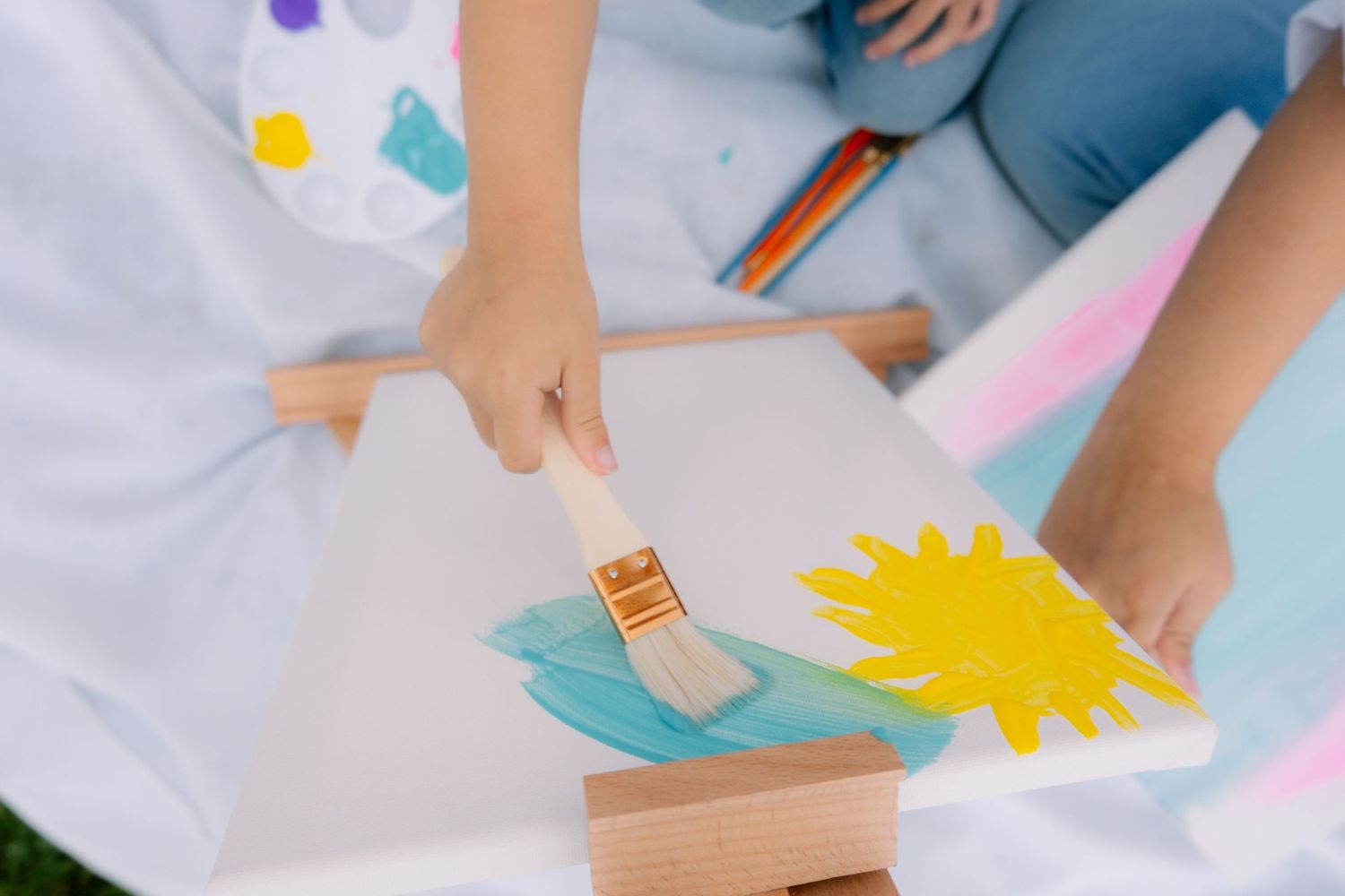 Children painting colorful artwork on canvas with a paintbrush, wearing white smocks during an art class activity.